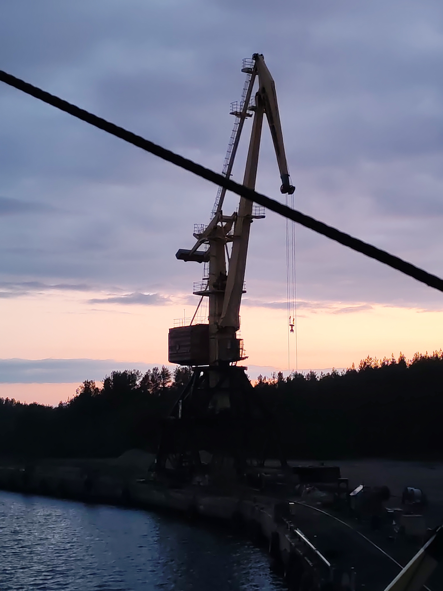 The image shows a port crane in Shalsky, Russia at sunset. The crane is beige, has a ladder attached to its side and a hook suspended on cables. It sits on a pier. A black rope diagonally bisects the image in the foreground. A line of trees is in the background silhouetted against the pink and gray sky. Calm water is visible to the left of the pier.