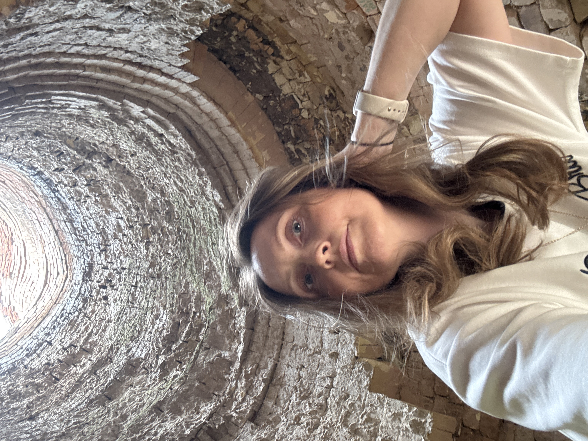 The media file features a young woman taking a low-angle selfie inside a circular brick structure. She is wearing a white t-shirt and a light-colored watch on her left wrist. Her long, light brown hair is draped over her shoulder and she is smiling gently at the camera. The background consists of the interior of what appears to be a kiln or a similar industrial structure, with rough, aged brickwork curving upwards and inwards. The lighting suggests it is daytime, with light coming from an opening at the top of the structure. The overall mood is relaxed and possibly adventurous, given the unusual perspective and setting. There is no visible text in the image. The location context provided is Mezhevoy, Russia.