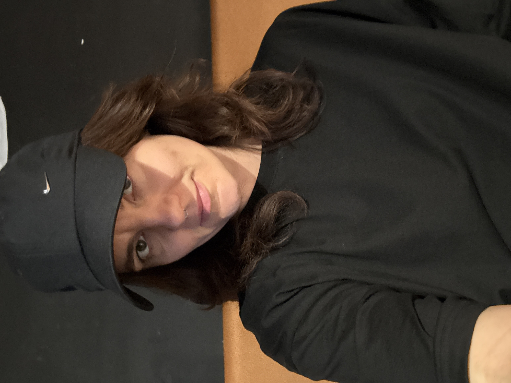 A woman wearing a black baseball cap with a white Nike logo and a black shirt is posing for a picture. She has brown hair and is looking at the camera with a slight smile. A brown surface is partially visible in the background. The location is not discernible from the image.