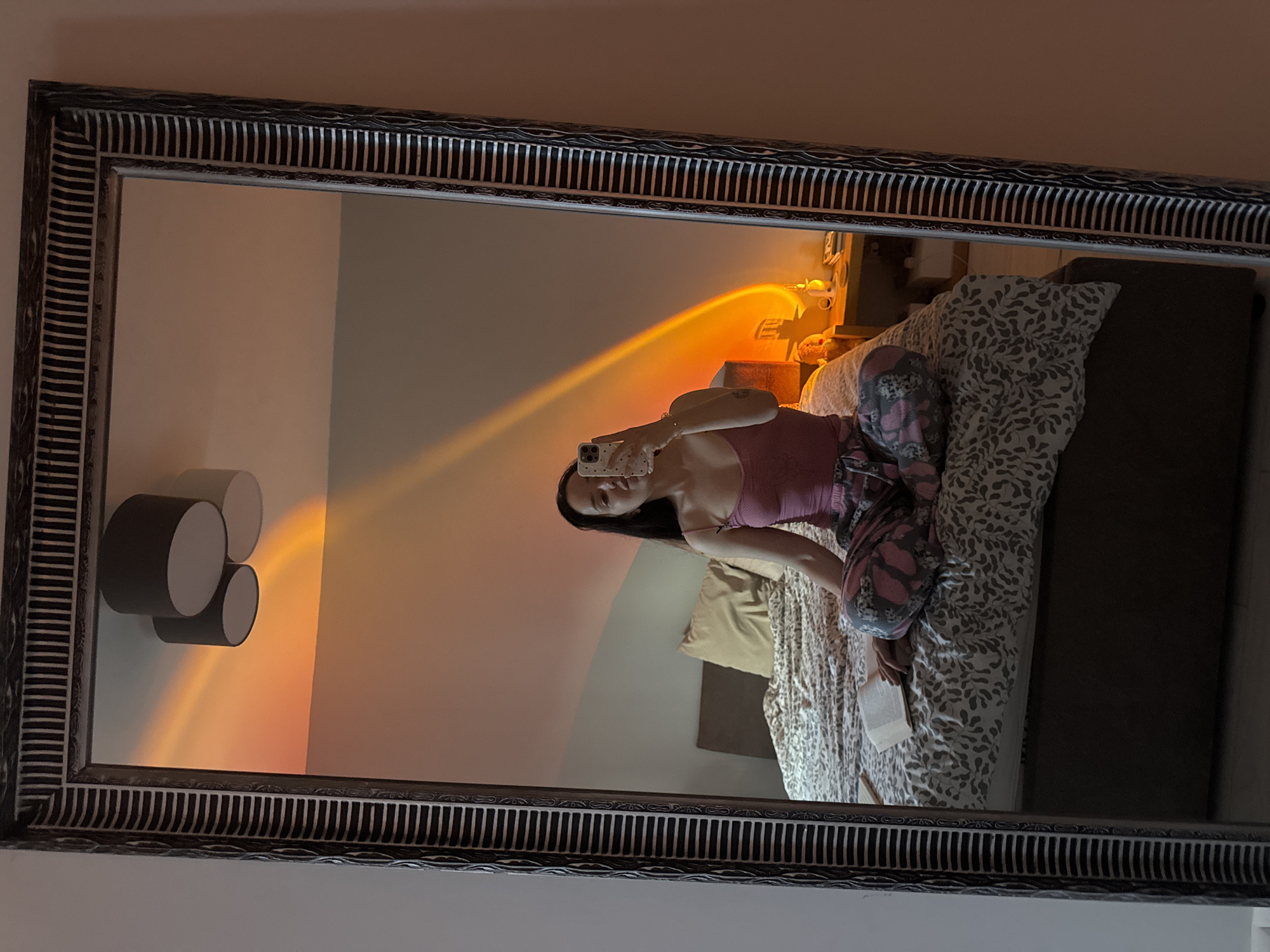 A mirror selfie captures a person in a bedroom setting. The large, ornate mirror, framed with a dark, textured border featuring horizontal stripes, reflects the scene. In the reflection, a person with long, dark hair is seated on a bed, holding a smartphone with a light-colored, patterned case up to take the photograph. She is dressed in a pink or mauve tank top and patterned pajama bottoms, which are grey with a distinctive floral or leaf design in pink and black.
The bed is adorned with patterned bedding, displaying a grey and white leaf or floral motif, with a light brown pillow resting against the headboard. The person appears to be holding a small book or notepad in her lap. To the right of the bed, a dark nightstand supports a lamp emitting a strong, warm orange and yellow light, casting a vibrant, diagonal beam onto the wall, characteristic of a "sunset lamp." On the opposite wall, to the left of the bed in the reflection, there is a modern wall fixture composed of three circular elements, two dark and one light.
The overall lighting suggests evening or nighttime, with the warm artificial light creating a cozy and atmospheric glow. The person's expression is neutral, focused on taking the picture. No discernible text is visible on the phone screen or other objects. The scene depicts a relaxed, personal moment within a bedroom.