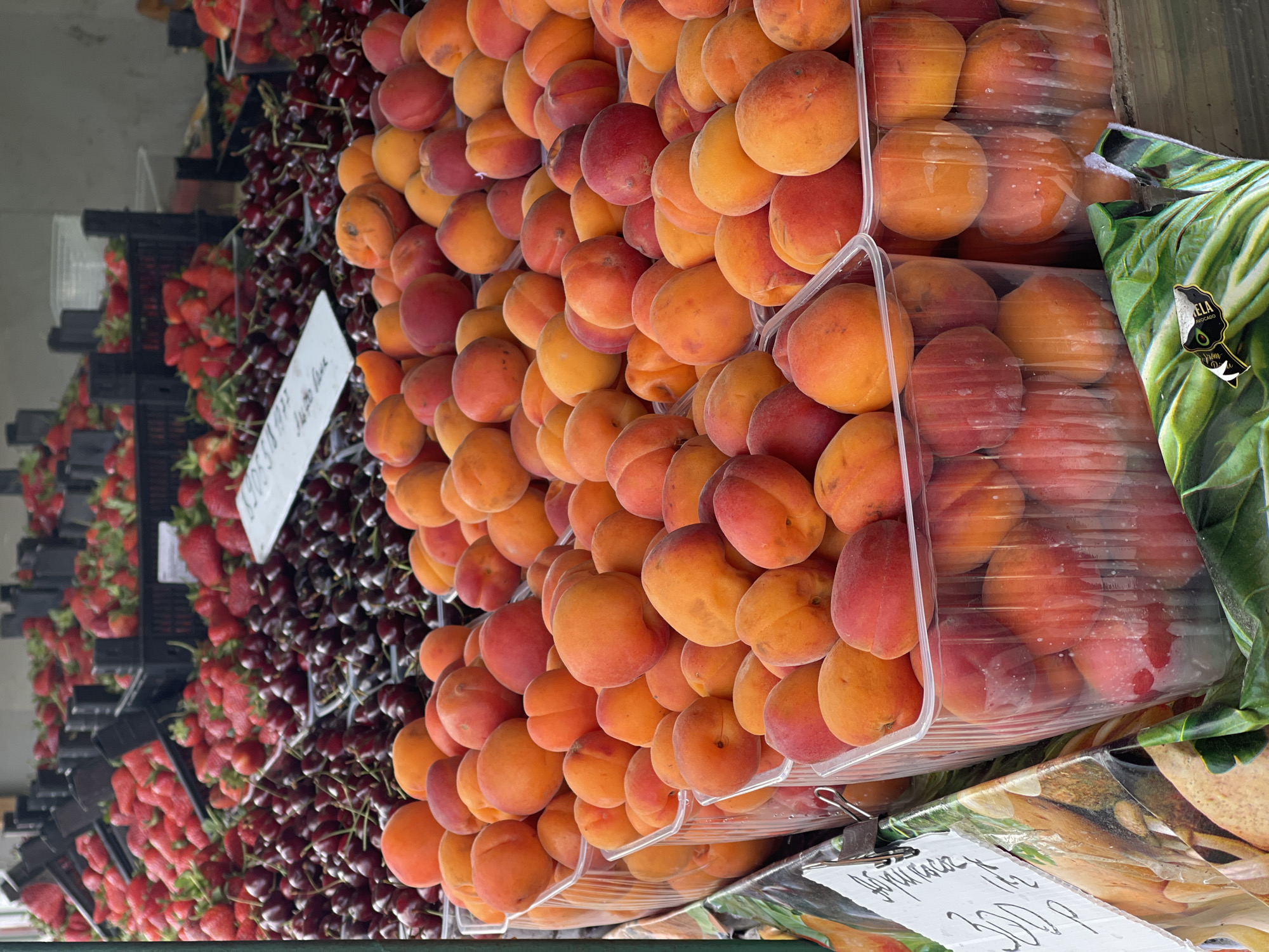 The image shows a fruit stand at an outdoor market, likely in Unknown, Unknown. The stand displays strawberries in black plastic crates on the left, followed by cherries. Next to the cherries are neatly stacked containers of ripe apricots, with some containers sealed in plastic wrap. A sign with the price of the apricots (300p) is visible. A bag of Avocados is visible on the right of the fruit stand.