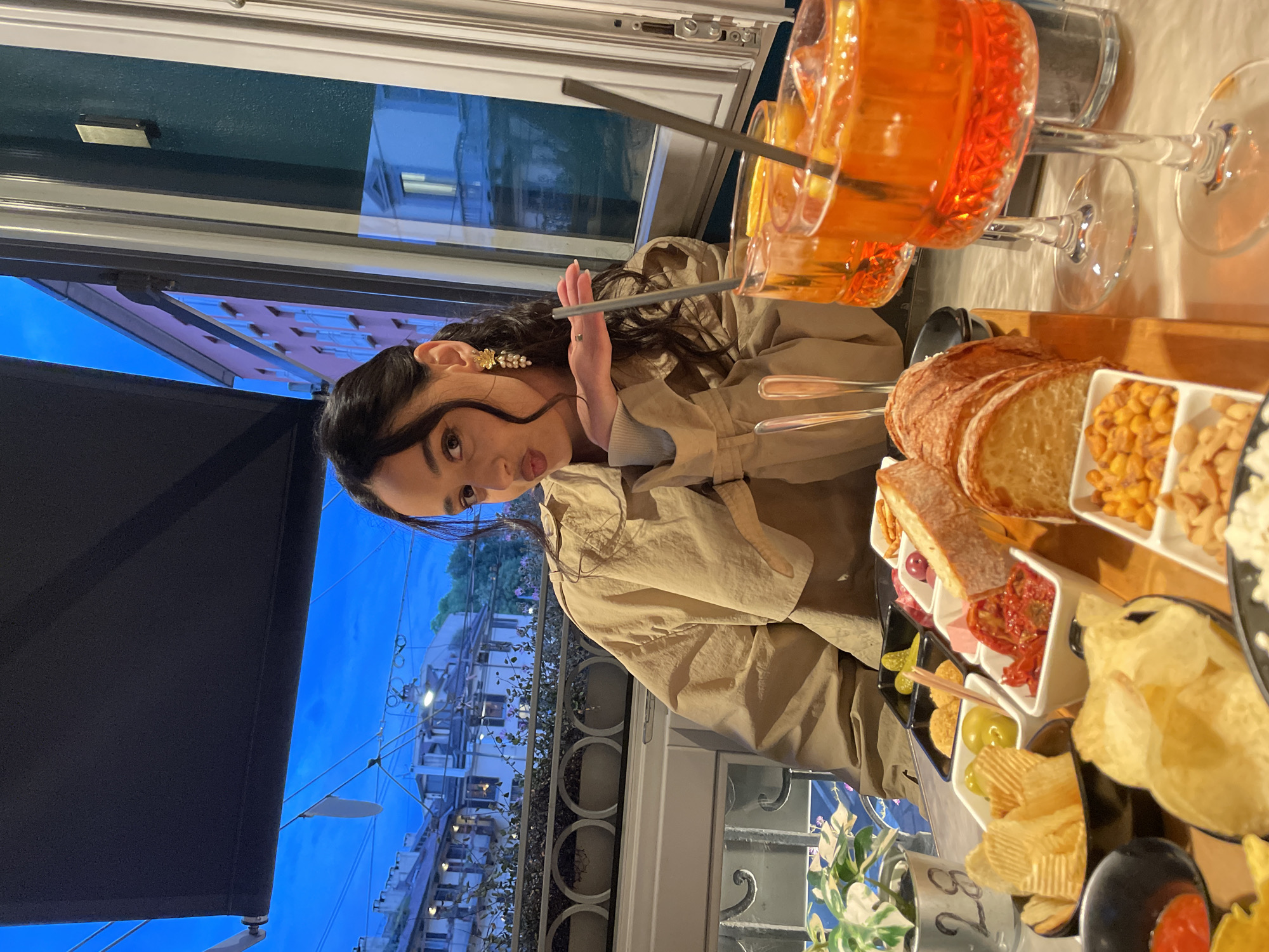 A woman with dark hair is seated at a table on a balcony or terrace overlooking a street in Milan, Italy, likely during dusk or evening. She is wearing a beige jacket and accessorized with floral-shaped earrings, and she is holding a drinking straw near her face with her hand.
On the table, there's a large glass of an orange-colored cocktail, possibly an Aperol Spritz, with a straw in it. Next to the glass is a wooden board filled with various appetizers, including slices of bread, nuts, chips, olives, pickles, sun-dried tomatoes, and other unidentified snacks served in small white bowls.
In the background, the cityscape of Milan is visible, featuring buildings and streetlights. The wrought-iron railing of the balcony is also visible, and a partial awning or shade is extending outwards, framing the scene. The lighting suggests it's either late afternoon or evening, with the sky a deep blue color.