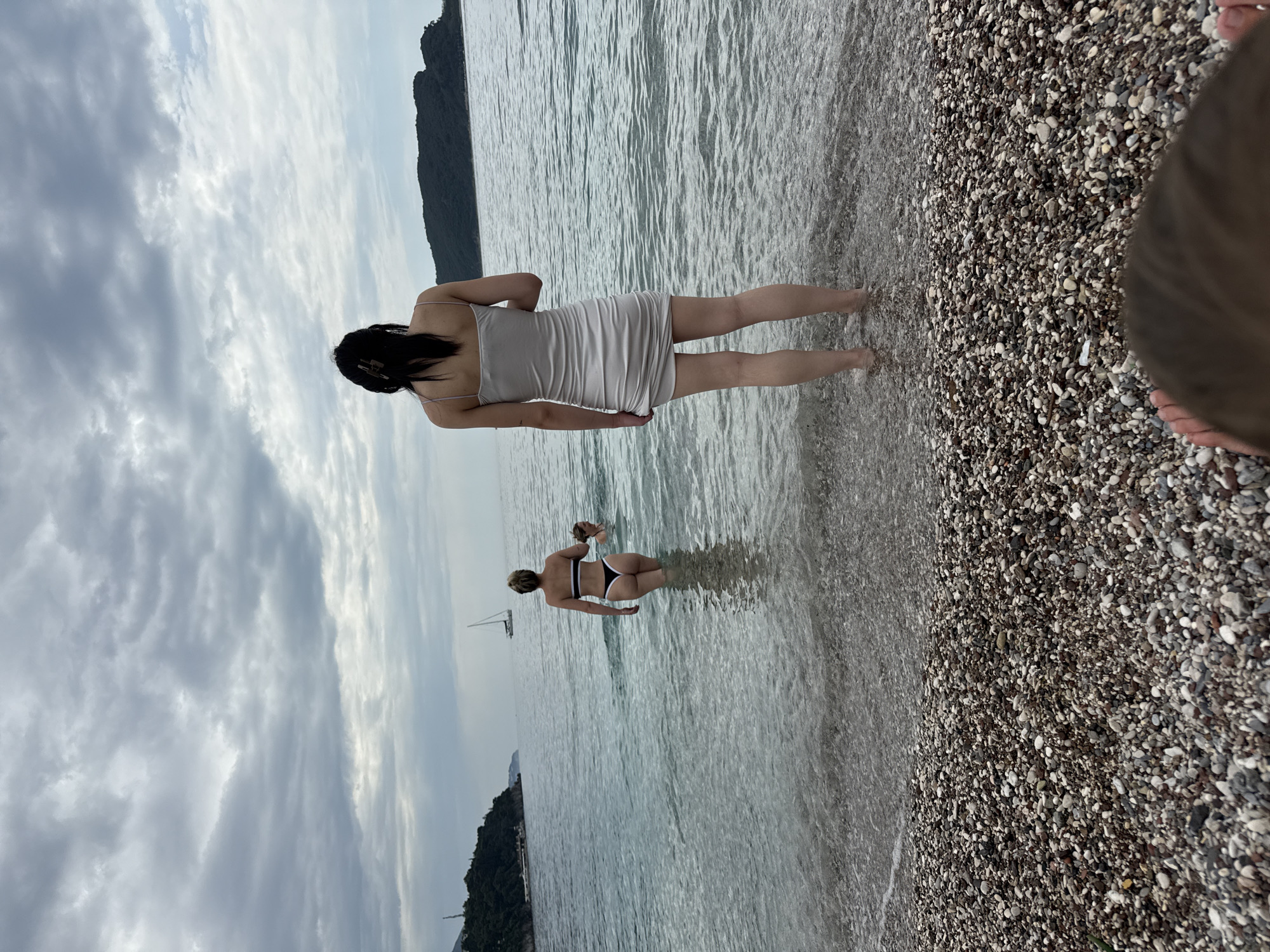 The image captures a scene on a pebble beach, likely in Budva, Montenegro, given the coastal environment and distant landmasses. A woman with dark hair, wearing a light-colored dress, stands near the shoreline, partially submerged in the water. Further into the water, another woman in a black and white bikini wades. A sailboat is faintly visible in the distance. The sky is overcast with grey clouds. The beach is composed of small, multicolored pebbles, and a portion of another person's head is visible on the right.
