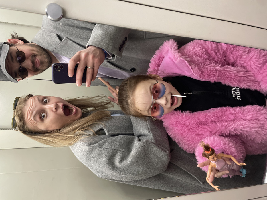 This is a mirror selfie taken indoors, presumably in Moscow, Russia. The scene features three individuals: an adult woman, an adult man, and a young girl, all visible from the waist up.
The woman, positioned on the left, has blonde hair and wears sunglasses pushed onto her head. She is wearing a grey overcoat and has an exaggerated expression with wide eyes and an open mouth. Her right hand is partially visible, holding a light-colored, slender stick-like object.
The man, on the right, has dark hair, a beard, and wears a grey baseball cap along with dark sunglasses. He is also wearing a grey overcoat over a light purple shirt. He holds a dark-colored smartphone in his right hand, reflecting a wedding band on his ring finger, indicating he is taking the selfie. He has a slight smile.
In the foreground, centered and below the adults, is a young girl with brown hair. She wears pink and blue aviator-style sunglasses and a bright pink faux-fur jacket over a black top. A white lollipop is visible in her mouth, and she also displays a wide-eyed, open-mouthed expression similar to the woman. Her left hand is holding a Barbie doll, dressed in a light purple outfit and blue shoes. The text on her black top is reversed due to the mirror reflection.
The environment appears to be an indoor space with plain, light-grey paneled walls, possibly an elevator or a fitting room, as indicated by the reflective surface and minimalist background. A silver metallic fixture is partially visible on the right side of the mirror.
