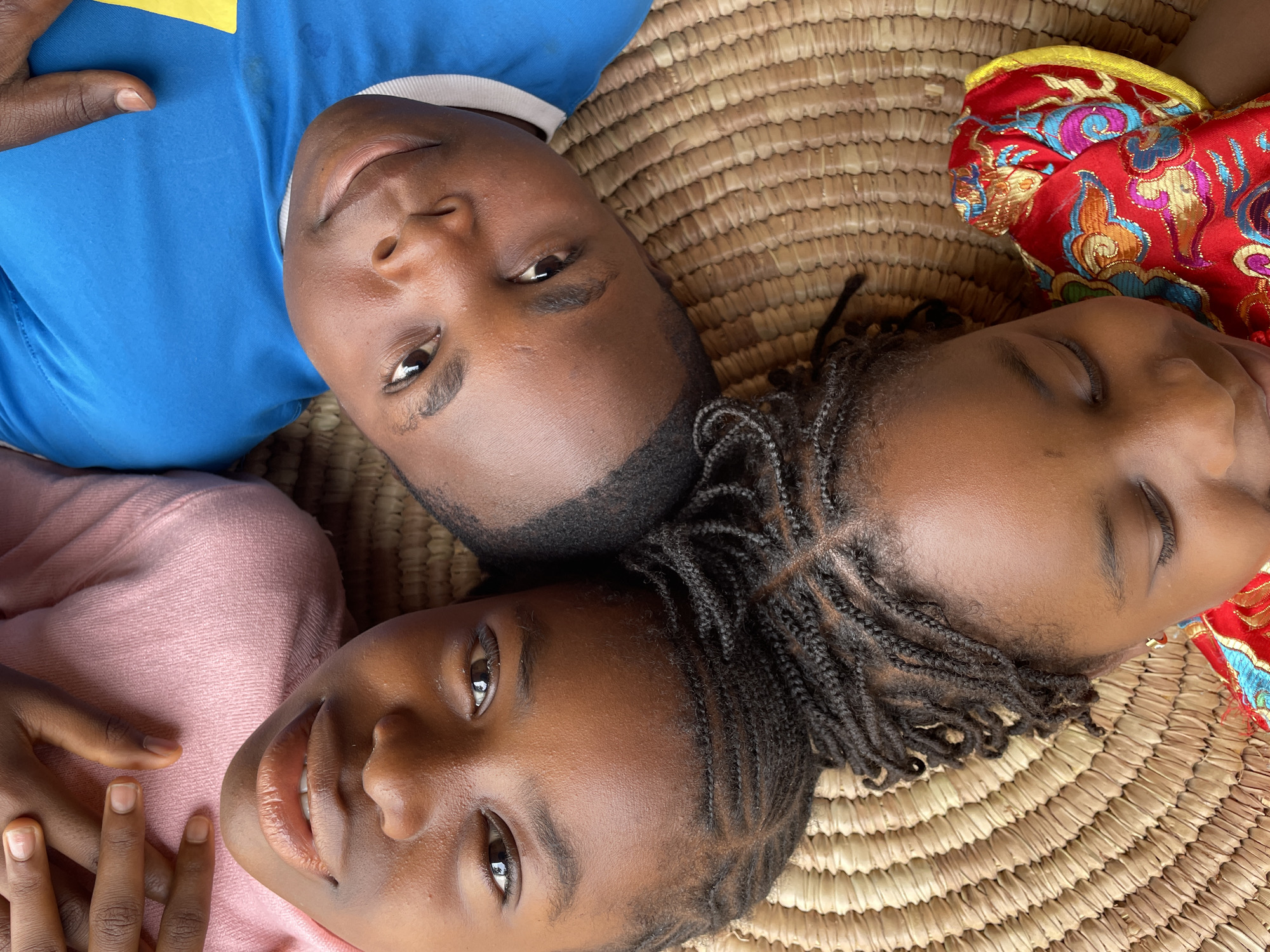 Three Black children are lying head-to-head on a woven mat. One child wears a blue shirt with a yellow patch and looks at the camera. The second child wears a pink shirt and also looks at the camera with a smile. The third child wears a red and gold ornate garment and has their eyes closed. The scene is located in Jos, Nigeria, as inferred from the subjects' ethnicity and attire.
