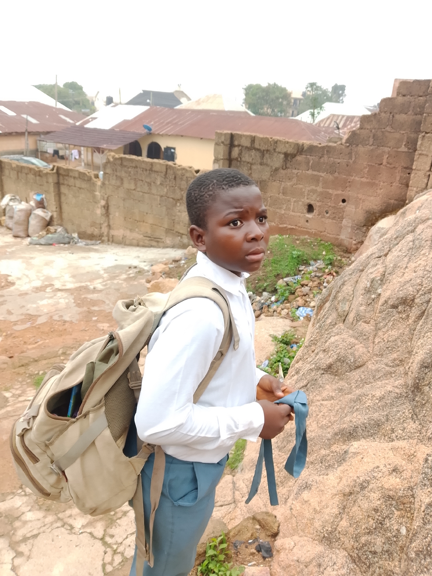 A young Black boy, appearing to be of school age, stands on a rocky incline. He is wearing a white collared shirt and blue trousers, and a tan backpack is slung over his shoulders. In his hands, he holds a blue tie, which he appears to be adjusting. The background reveals a village scene with buildings topped with corrugated metal roofs, a stone wall, and sacks piled near the wall. The ground is a mix of dirt and patchy vegetation, with some scattered debris. The lighting suggests an overcast or hazy day.