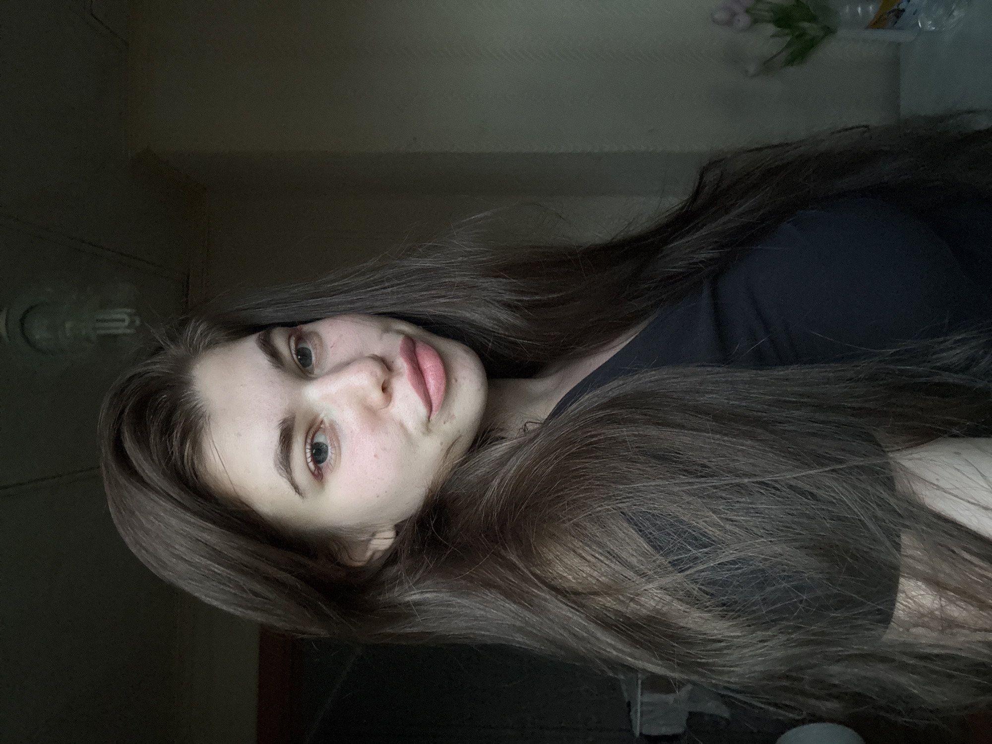 A young woman with long, brown hair and fair skin is facing the camera. She has light-colored eyes and pink lips. She is wearing a black t-shirt. The background is dimly lit and features a wall, a light fixture, and a vase of flowers. The location, Мещёры, Russia, is not discernible from the image.