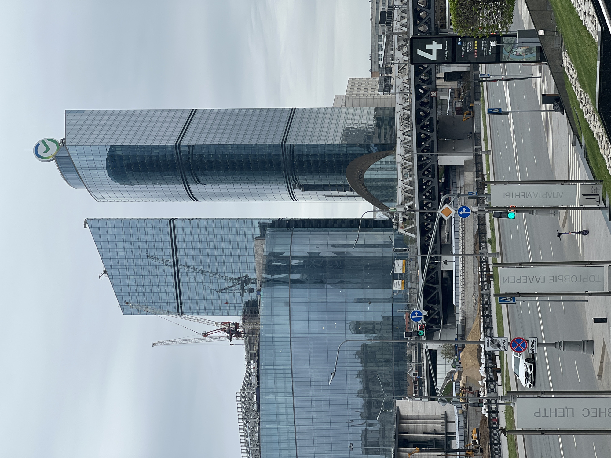 The image captures a view of the Moscow International Business Center, also known as "Moscow City," in Russia. The scene is oriented vertically, with the skyscrapers towering upwards. The buildings are primarily glass and steel structures, reflecting the sky and surrounding environment. In the foreground, there is a roadway with street signs in Russian, indicating an urban setting. A few construction cranes are visible, suggesting ongoing development in the area. The sky is overcast, casting a diffused light across the scene. There is no evidence of human activity or people. A white vehicle is seen parked on the side of the road.