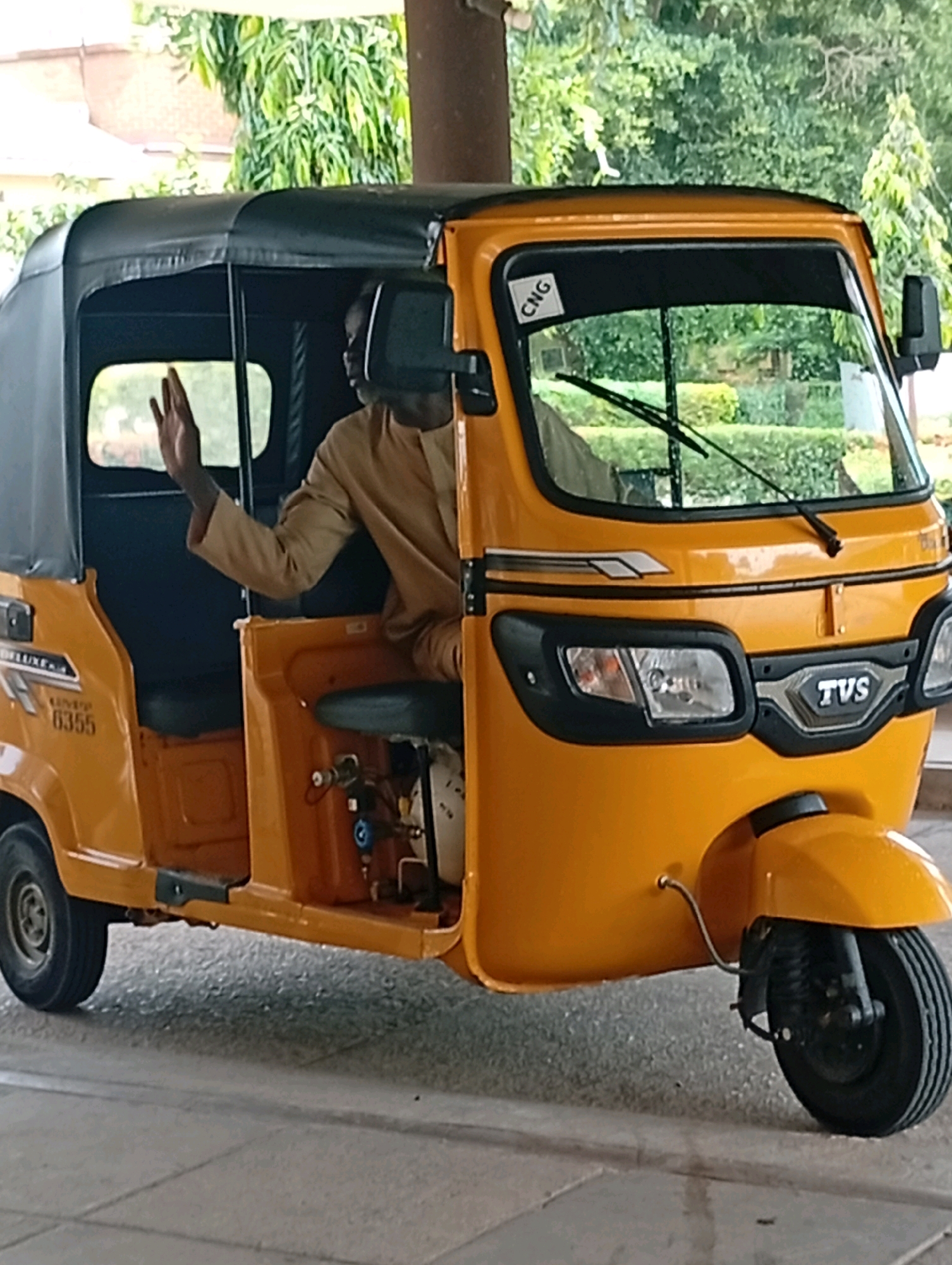 A driver in a tan-colored garment is seated inside a yellow three-wheeled auto rickshaw, also known as a Keke Napep. The driver's left hand is raised, palm facing forward, in a gesture that could be a wave or signaling. The auto rickshaw has a black fabric canopy covering the passenger area. A "CNG" sticker is visible on the windshield. The vehicle displays the brand "TVS" on its front grille and has a model number "6355" and "DELUXE" branding on its side. The auto rickshaw is parked on a paved surface with visible grout lines, suggesting an outdoor or semi-covered urban setting. The background shows green foliage and trees, indicating a potentially green or park-like area within the city of Gombe, Nigeria. The overall scene appears to be a moment captured in a typical urban environment of Gombe.