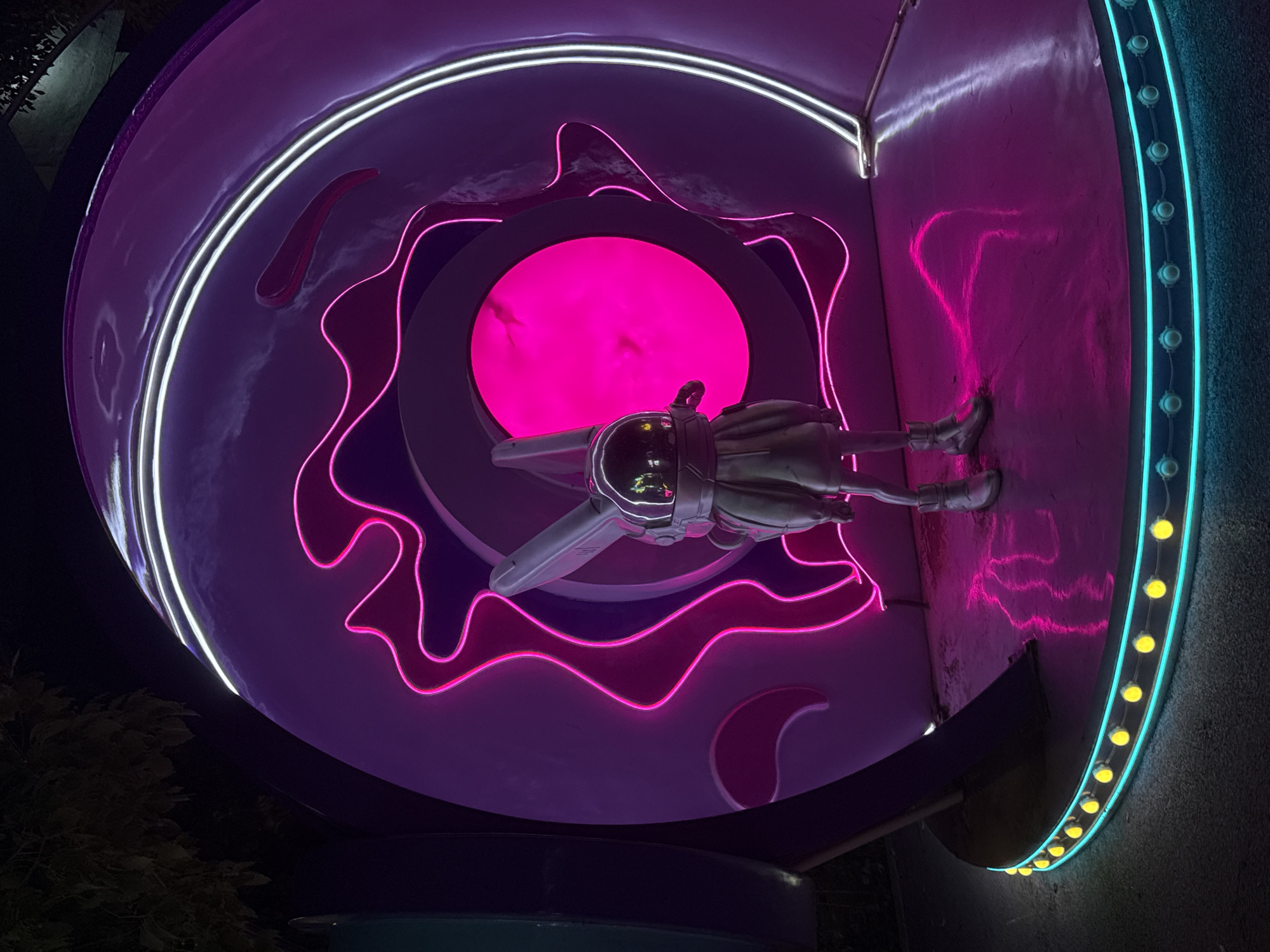 The image depicts a nighttime outdoor scene in Yiwu, China, likely an art installation or a themed attraction. In the center stands a solitary figure, a statue or mannequin resembling a rabbit in an astronaut suit, complete with a helmet and large ears. The figure is positioned on a circular, reflective platform illuminated by a ring of yellow lights and a turquoise neon border.
Behind the figure is a large, circular structure with a striking purple exterior. This structure is adorned with bright pink and white neon lights that create swirling, organic shapes. The inner part of the circle features a vibrant, glowing pink orb, suggesting a portal or a central focal point. The overall aesthetic is futuristic and vibrant, with strong contrasts between light and shadow. The background is dark, with the suggestion of trees and foliage on the left, indicating an outdoor setting. The reflective surface of the platform mirrors the neon lights, enhancing the visual dynamism of the scene.