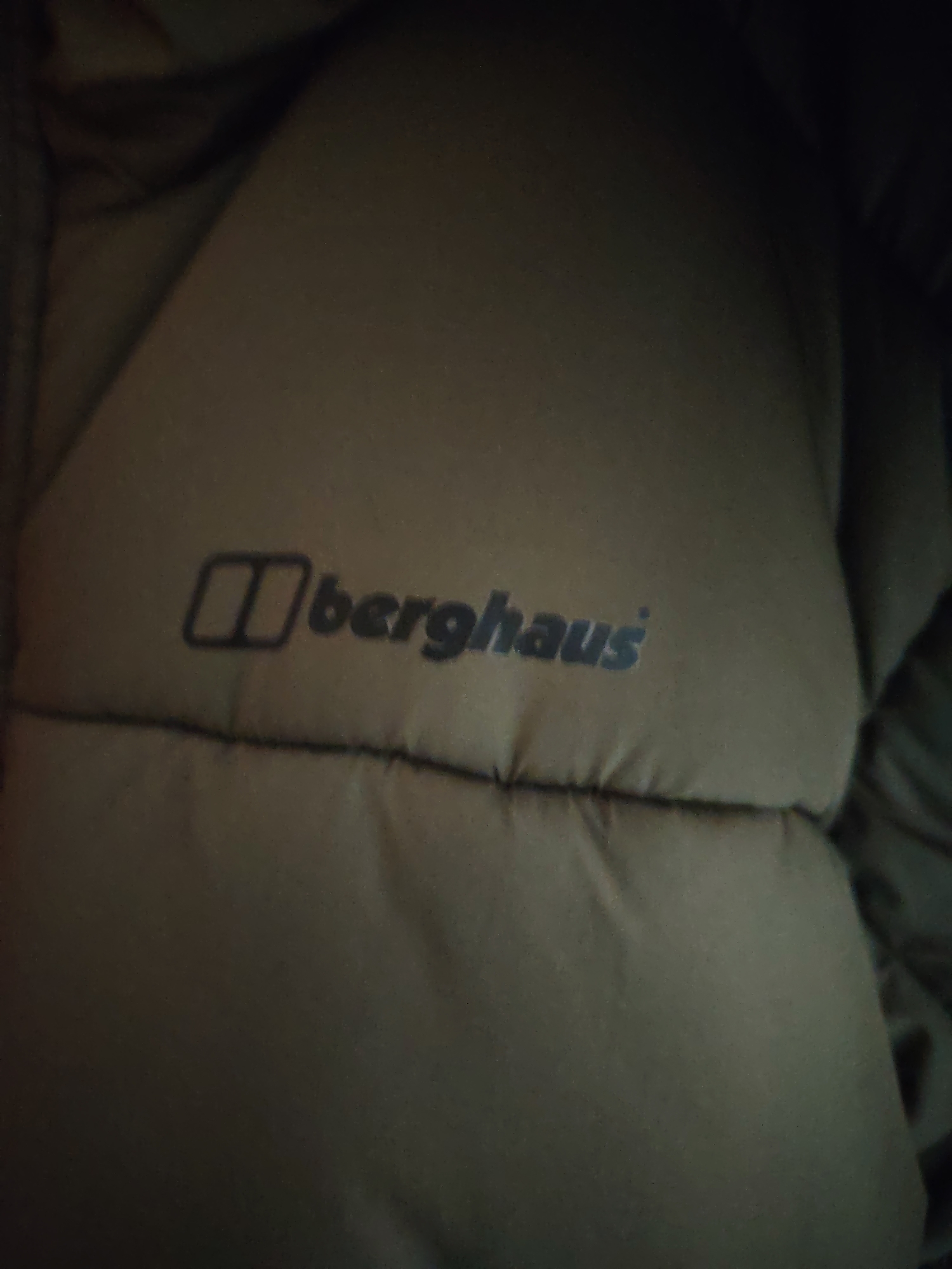The image presents a close-up, top-down view of a dark-colored, padded garment, likely a jacket or sleeping bag, against a dimly lit background. The fabric exhibits a muted olive-green or dark grey-brown hue. Prominently displayed in the upper-central area is the "Berghaus" brand logo, rendered in a darker blue-grey tone. The logo comprises a rectangular emblem with two internal vertical bars preceding the word "Berghaus." A horizontal seam is visible below the logo, indicating a quilted or insulated construction. The scene is devoid of any discernible individuals, actions, or additional objects beyond the textile item. The lighting is low and somewhat uneven. This depiction is situated in Huntingdonshire, United Kingdom.