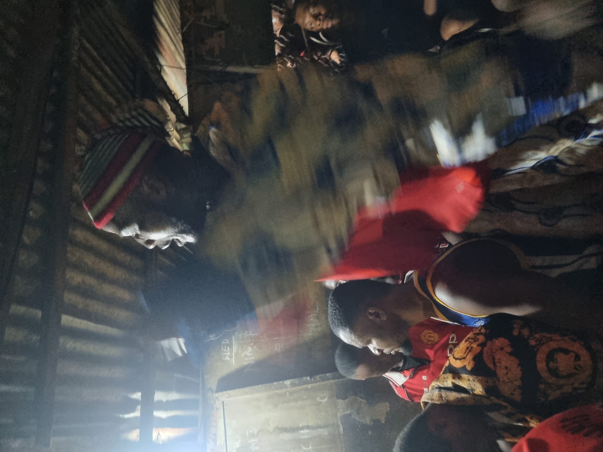 The image shows an indoor scene in what appears to be a room in Onitsha, Nigeria. Multiple people are visible, some with dark complexions. An older man is wearing a striped hat. Several other people, including a few young men, are wearing red shirts or clothing. One person is wearing a basketball jersey, while another is wearing a patterned shirt. The room has corrugated metal walls. The lighting is poor, with some bright spots obscuring parts of the scene.