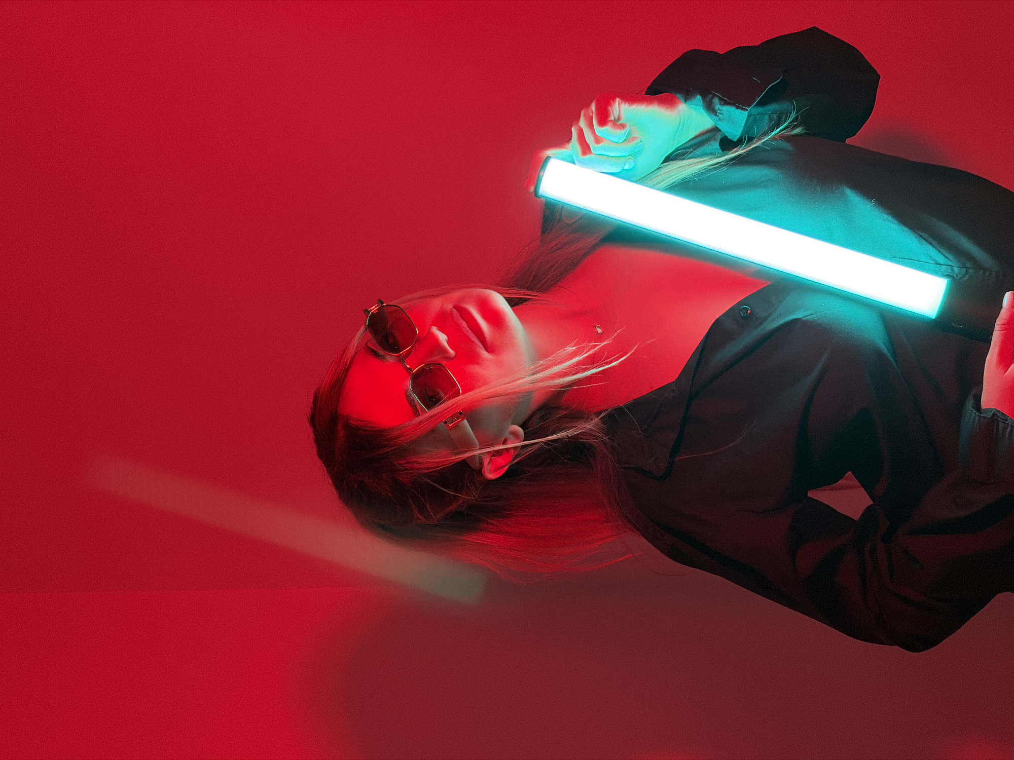 The image presents a stylized portrait of a person, likely a young woman, lying down and holding a bright, luminous tube light. The subject is oriented horizontally across the frame, with their head positioned towards the left. They have blonde hair that appears to be flowing slightly, and are wearing dark, reflective sunglasses and a black long-sleeved top or jacket.
Both hands of the subject firmly grip a glowing tube light, which emits a strong, cool blue/cyan light and extends diagonally across their upper body. The scene is bathed in a vibrant, uniform red light, creating a striking color contrast between the red background and the blue light emanating from the tube. This dual lighting effect highlights the subject's face, hands, and parts of their hair with a cool blue glow, while other areas are imbued with the warm red.
The overall atmosphere is modern, artistic, and evocative of neon aesthetics. The subject's expression is obscured by the sunglasses, lending an enigmatic quality to the portrait. The background is a solid color, suggesting a controlled studio environment or a room purposefully lit for such effects. There are no visible location cues within the image to specify Budva, Montenegro, nor is there any visible text. The activity depicted is a posed photographic session.