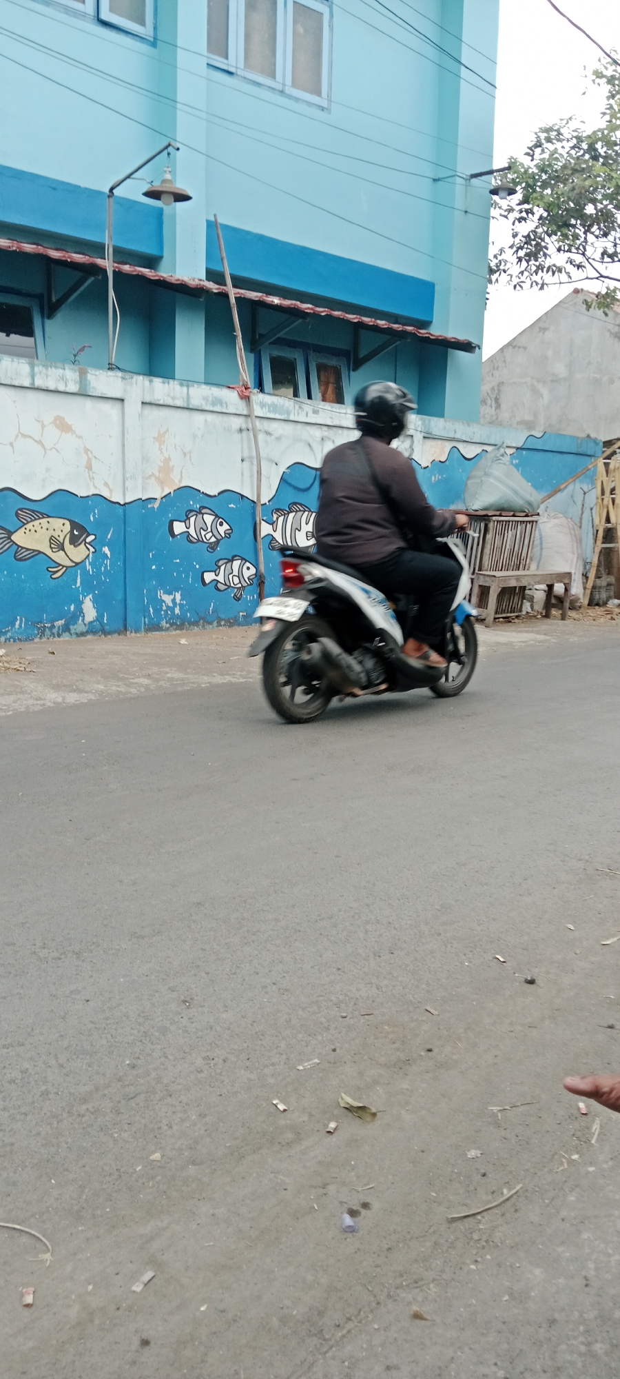 The image captures a street scene in Purwomartani, Indonesia, featuring a person riding a scooter. The scooter is white with blue accents and is moving from left to right across the frame. The rider is wearing a dark helmet and a dark long-sleeved shirt, with dark pants. The scooter's motion is blurred, indicating movement.
In the background, a light blue building with white-trimmed windows is visible. Below the building, a blue wall with a mural of stylized fish swimming in blue waves is painted. A street lamp extends from the building over the wall. To the right of the mural, a stack of items covered with a light-colored tarp and some wooden structures are present. The road surface is asphalt and appears somewhat worn, with some debris scattered on it, including small pieces of paper and a dried leaf. The sky is overcast.