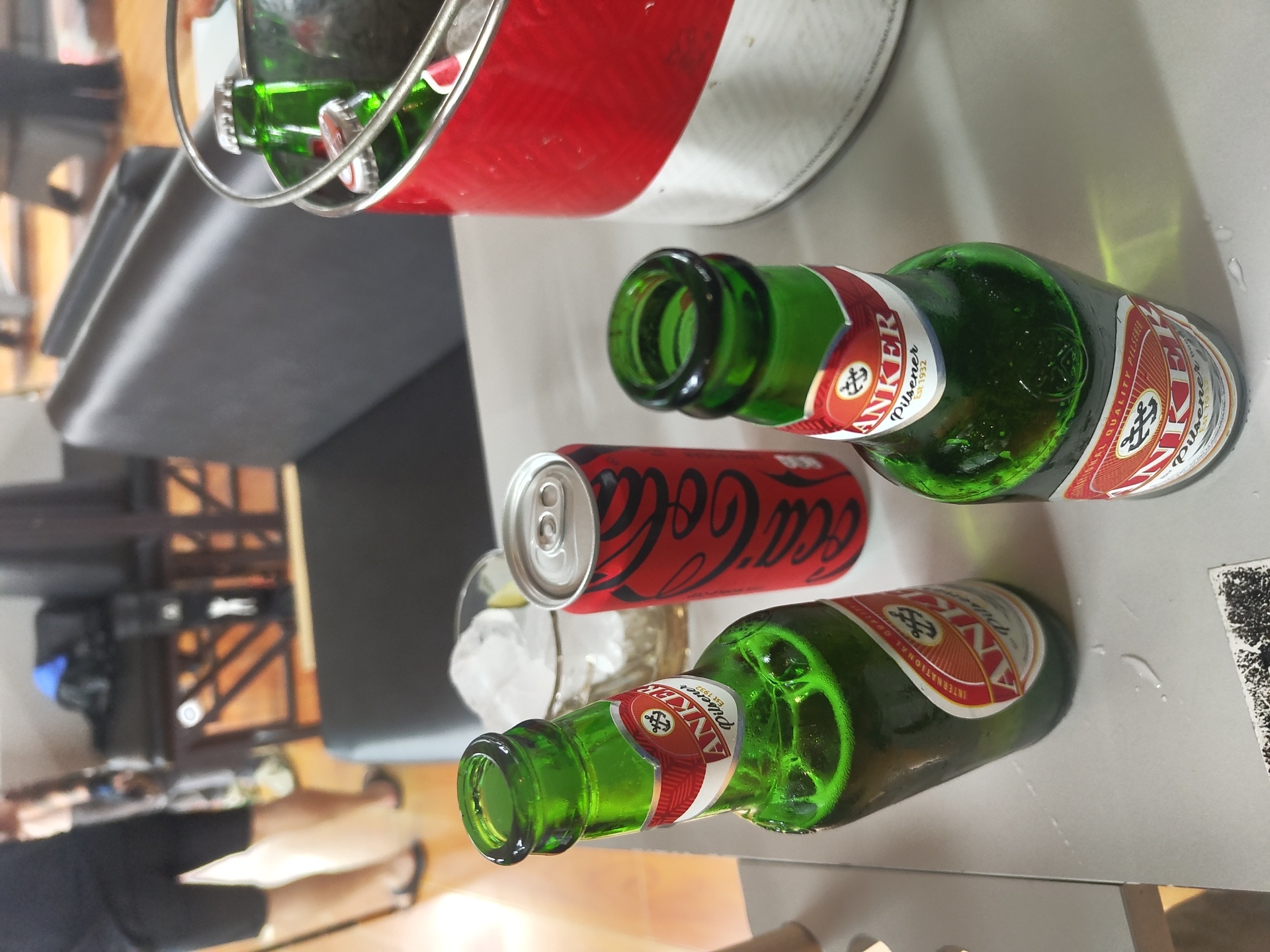 On a light grey table, there are two green glass bottles of Anker Pilsener beer and one red Coca-Cola can. The bottles are laid on their sides. One bottle has a glass with ice beside it. A red and white metal bucket with several bottles inside is in the upper left corner. In the background, a partial view of a person's legs and feet is visible, along with furniture including a grey sofa and a wooden chair. The brand on the bottle indicates this scene could be in Kuta Selatan, Indonesia.