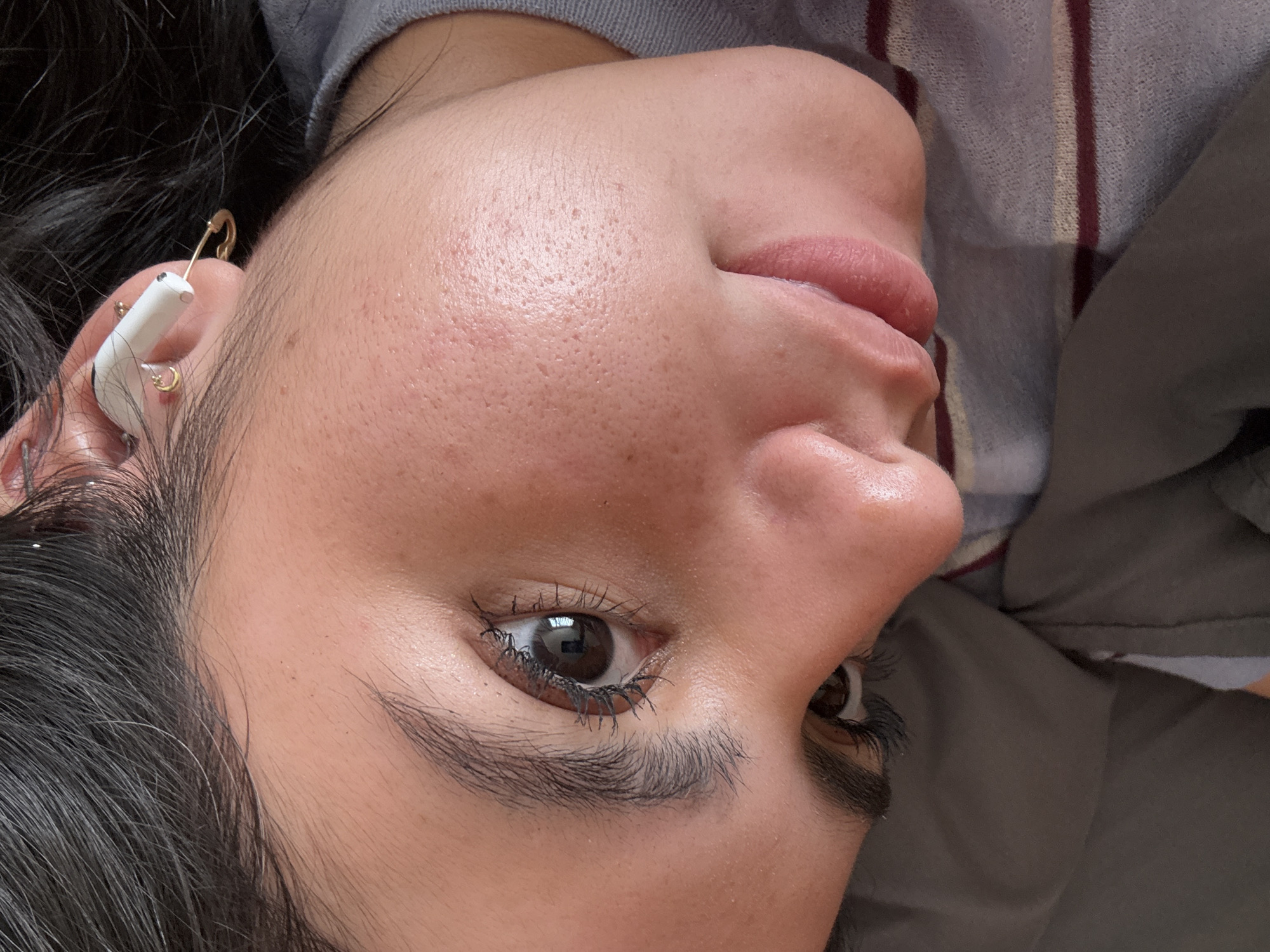 Close-up of a person's face, positioned at an angle. The individual has dark hair, brown eyes, and appears to have a white wireless earbud in their left ear. The earbud is decorated with a safety pin and small gold hoops. The person's skin texture is visible, with pores and minor blemishes present. The background is out of focus and appears to be a grey and striped garment. There is no indication in the image that can place the picture in the city of San Jerónimo Chicahualco, Mexico.