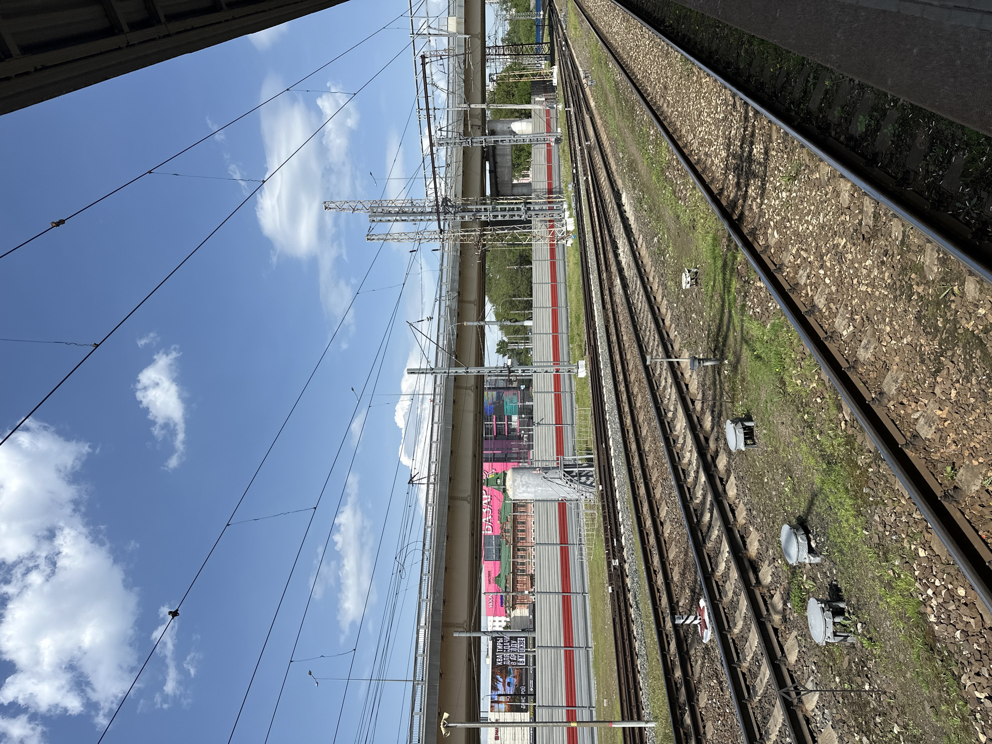 This image depicts a railway scene in Nizhny Novgorod, Russia. The view is from the edge of a platform, looking across several sets of railway tracks. To the left of the tracks is a concrete overpass structure adorned with colorful advertisement banners. Overhead power lines crisscross the sky above the tracks and the bridge. The tracks themselves are lined with ballast and interspersed with vegetation. Signal boxes are visible along the tracks. The sky is blue with scattered white clouds.