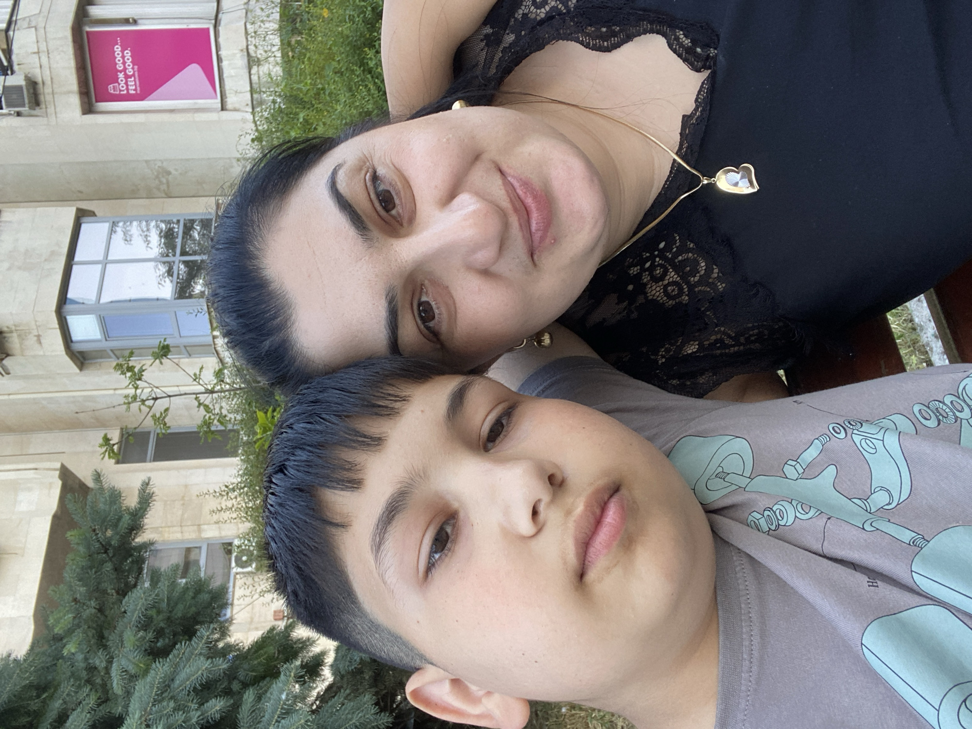 A medium shot of a woman and a young male child lying down together. The woman is older, with dark hair pulled back, dark eyes, and light skin. She is wearing a black lacy top and a gold necklace with a heart-shaped pendant. The child has dark hair, light skin, and is wearing a gray T-shirt with a blue design. They appear to be lying outside on a bench or grassy area near a building. In the background, there's greenery and a building with windows and a sign on the window that says "look good... feel good." The building is located in the city of Asenovgrad, Bulgaria.