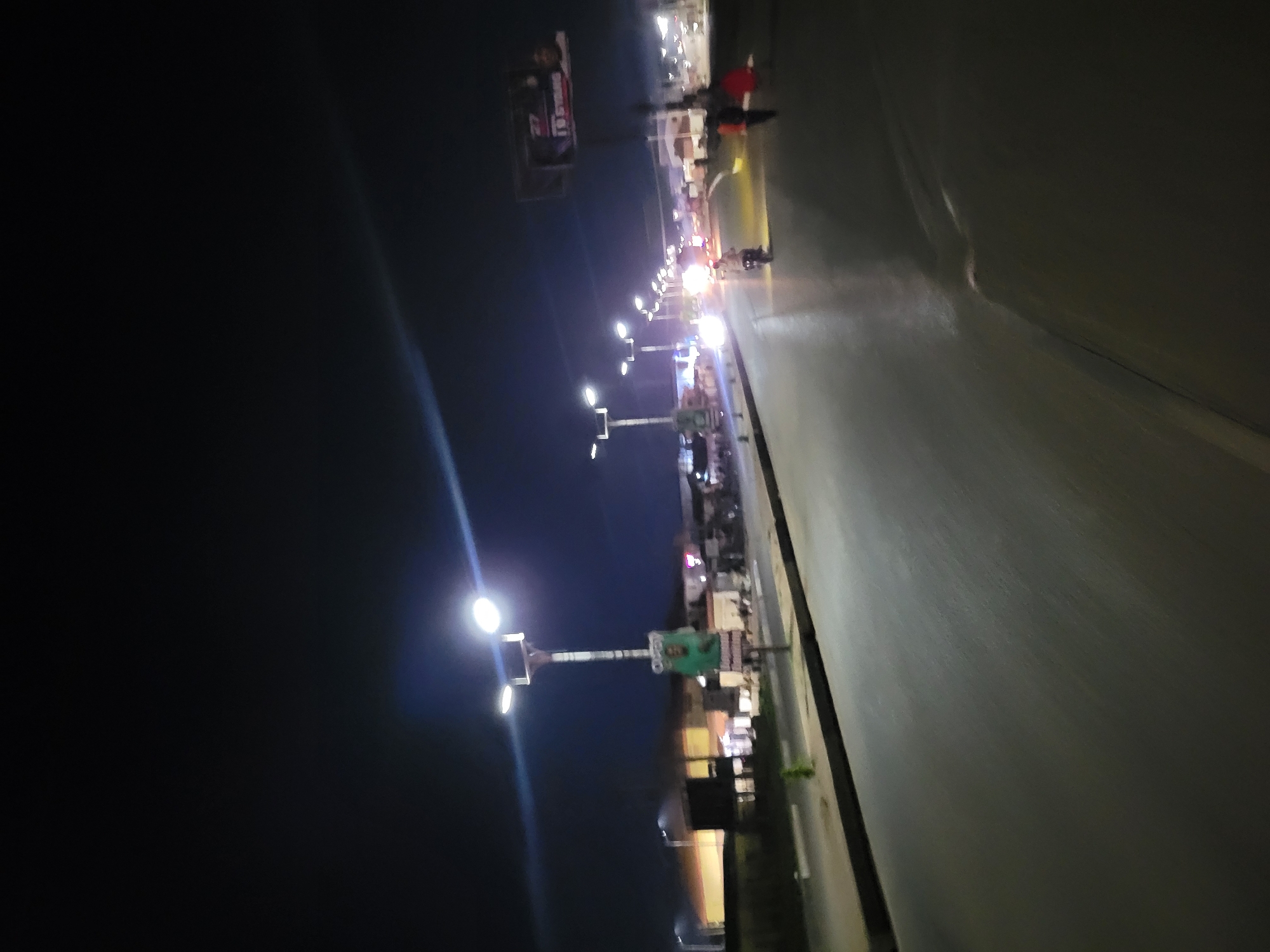 The image shows a street scene at night in an urban area. The view is along a road with buildings and streetlights lining one side. People and vehicles are visible on the road. The sky is dark.