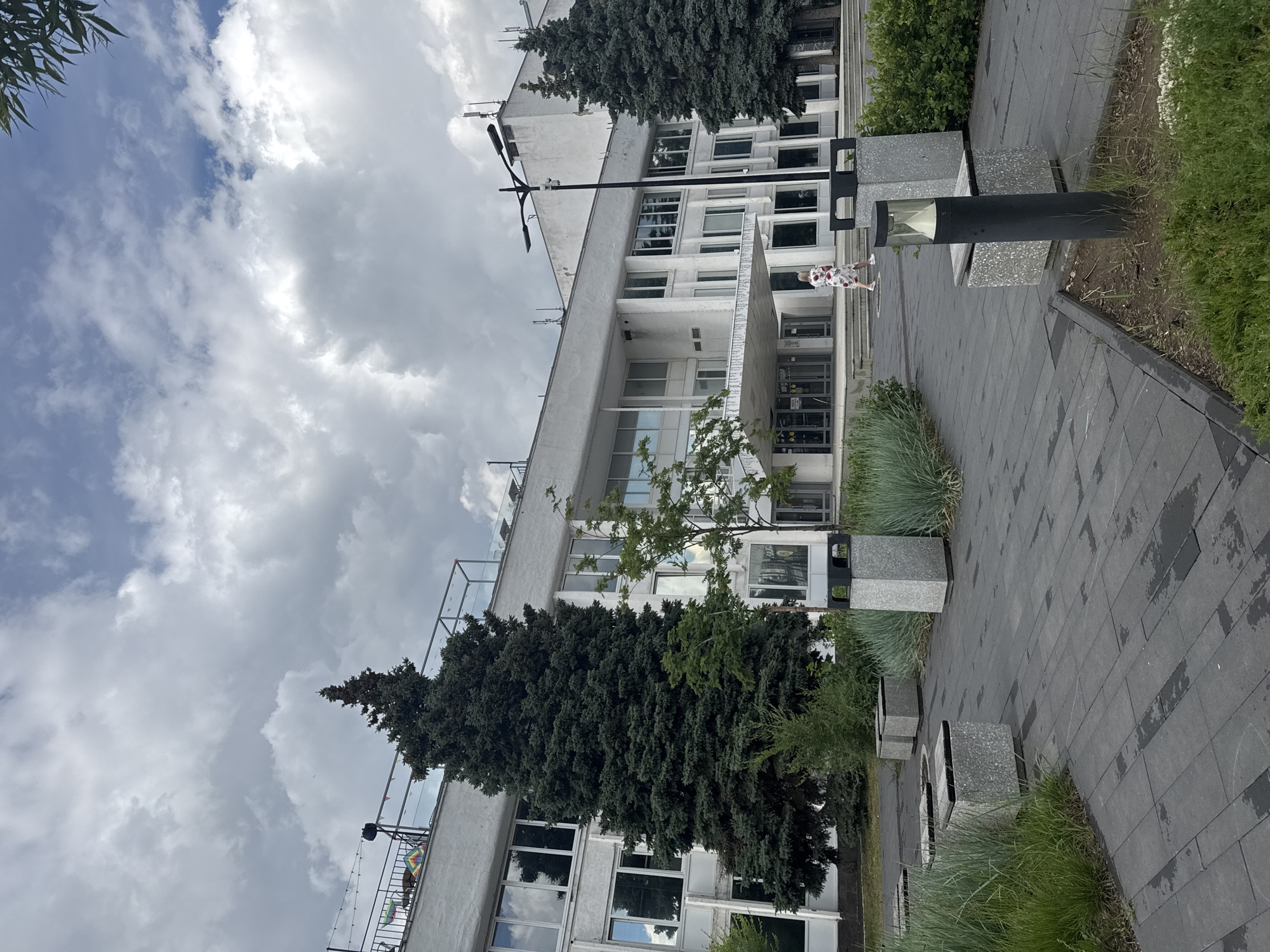This image depicts the exterior of a white, modernist building in Izhevsk, Russia. The building has a long, rectangular facade with numerous windows and a covered entrance. In front of the building, there is a paved walkway with several concrete benches and planters. Two tall evergreen trees stand on either side of the building's entrance. A single woman wearing a floral dress is walking up the steps towards the building. The sky is overcast with dramatic clouds. The overall impression is one of a public space or office building on a cloudy day.
