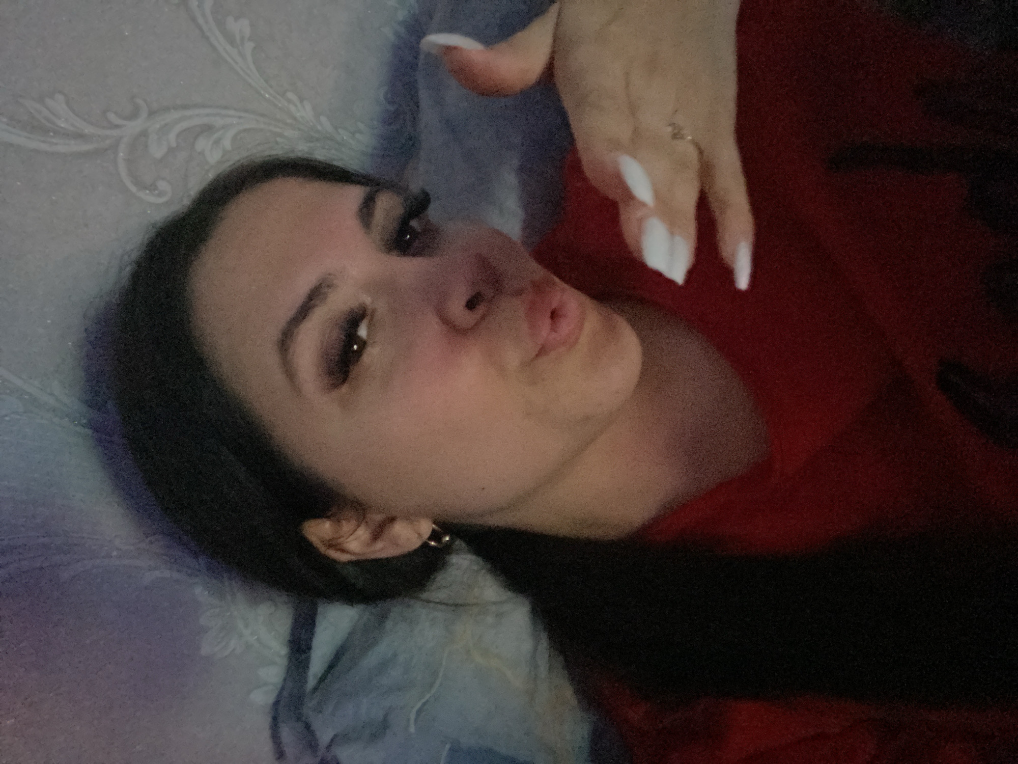 A woman is lying down, possibly in bed, on a blue and white patterned surface. She is wearing a red top. Her hair is dark and she has makeup on, including false eyelashes. Her fingernails are long and white. She is making a kissing face toward the camera. The location is not determinable from the image, and the mention of Chaikovsky, Russia, cannot be verified.