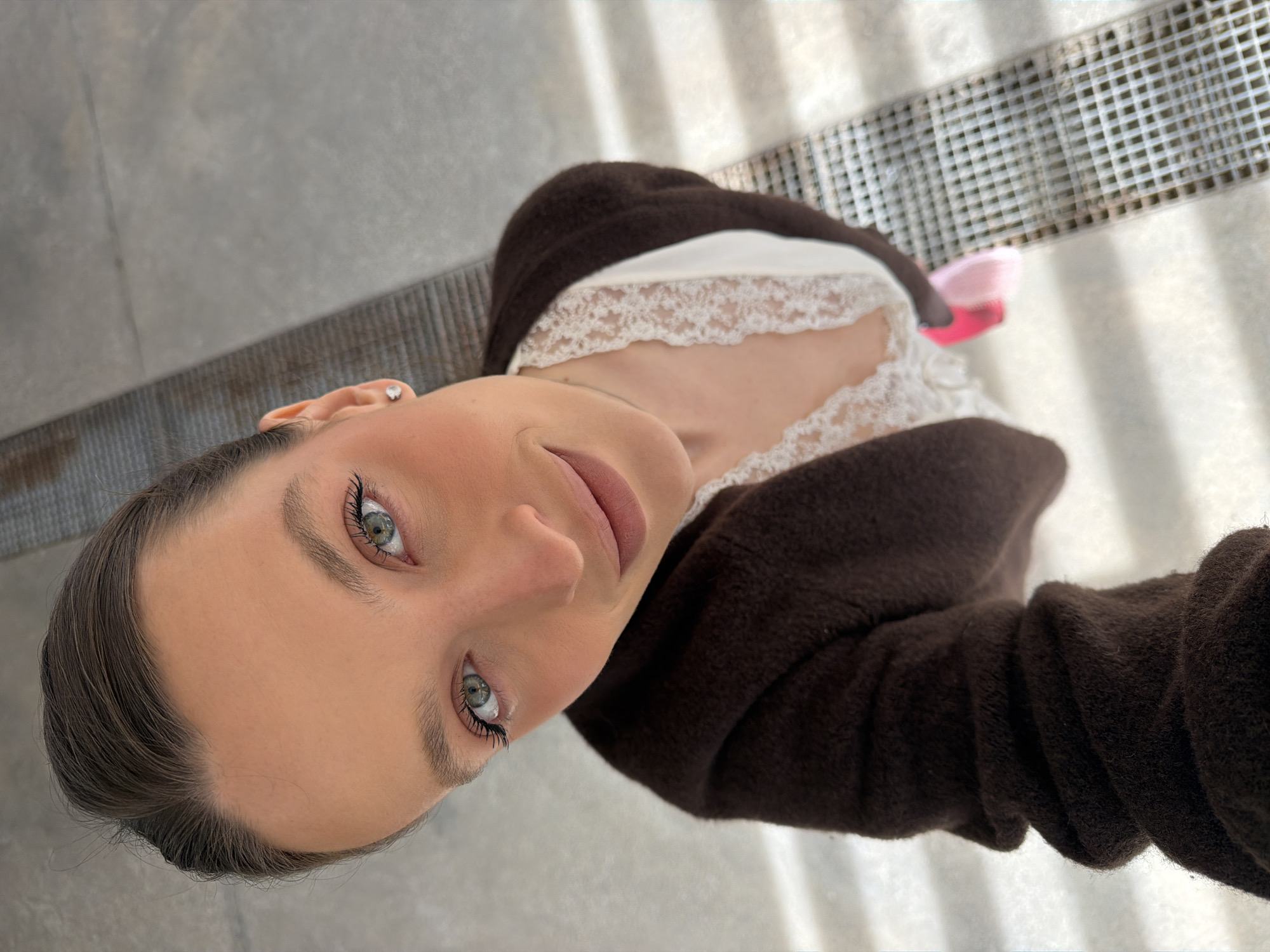 A woman with fair skin, light green eyes, and dark hair is facing the camera in a selfie. She is wearing a brown, textured cardigan over a white top with lace detailing around the neckline. She has a stud earring in her left ear. The background consists of a gray, concrete surface and a metal grate with linear patterns. The setting could be an outdoor area in Budva, Montenegro, but it is not readily identifiable from the image.