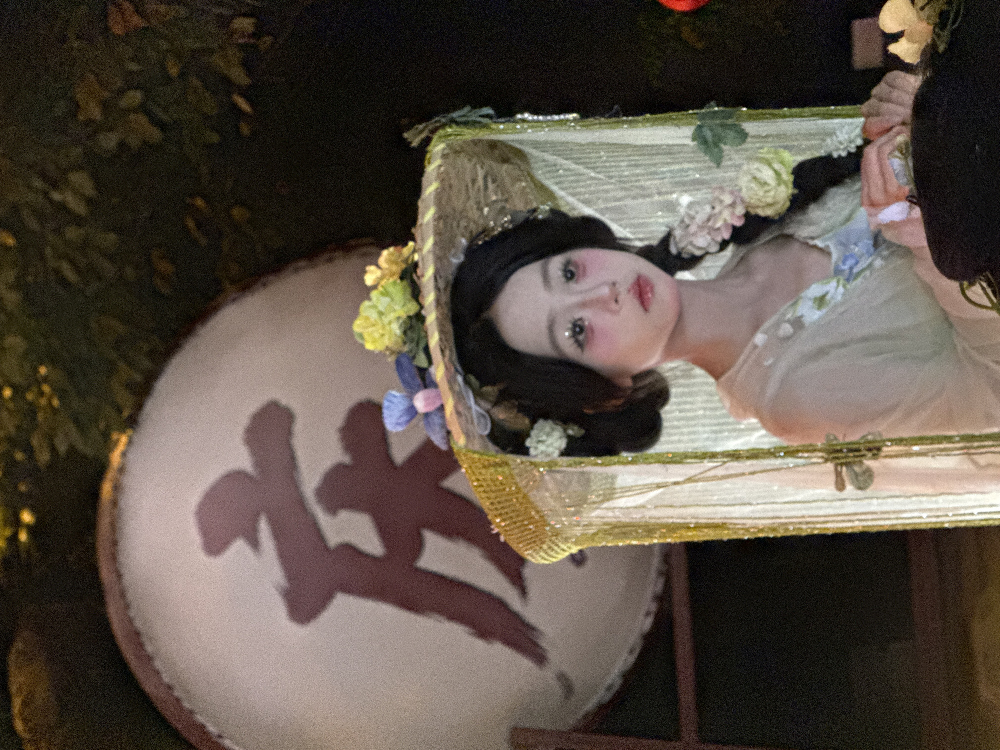 An Asian female wearing a traditional costume with a large, ornate hat adorned with flowers and a veil stands in front of a large drum with Chinese calligraphy. The female has her hair braided and wears makeup that accentuates her eyes and cheeks. She appears to be participating in a cultural event or performance. The background suggests an outdoor nighttime setting, possibly in Yuzhong District, China, indicated by the presence of traditional elements and the potential for a festival or celebration. The lighting is dim, with the focus on the woman and the drum, highlighting the artistic details of the costume and the calligraphy.