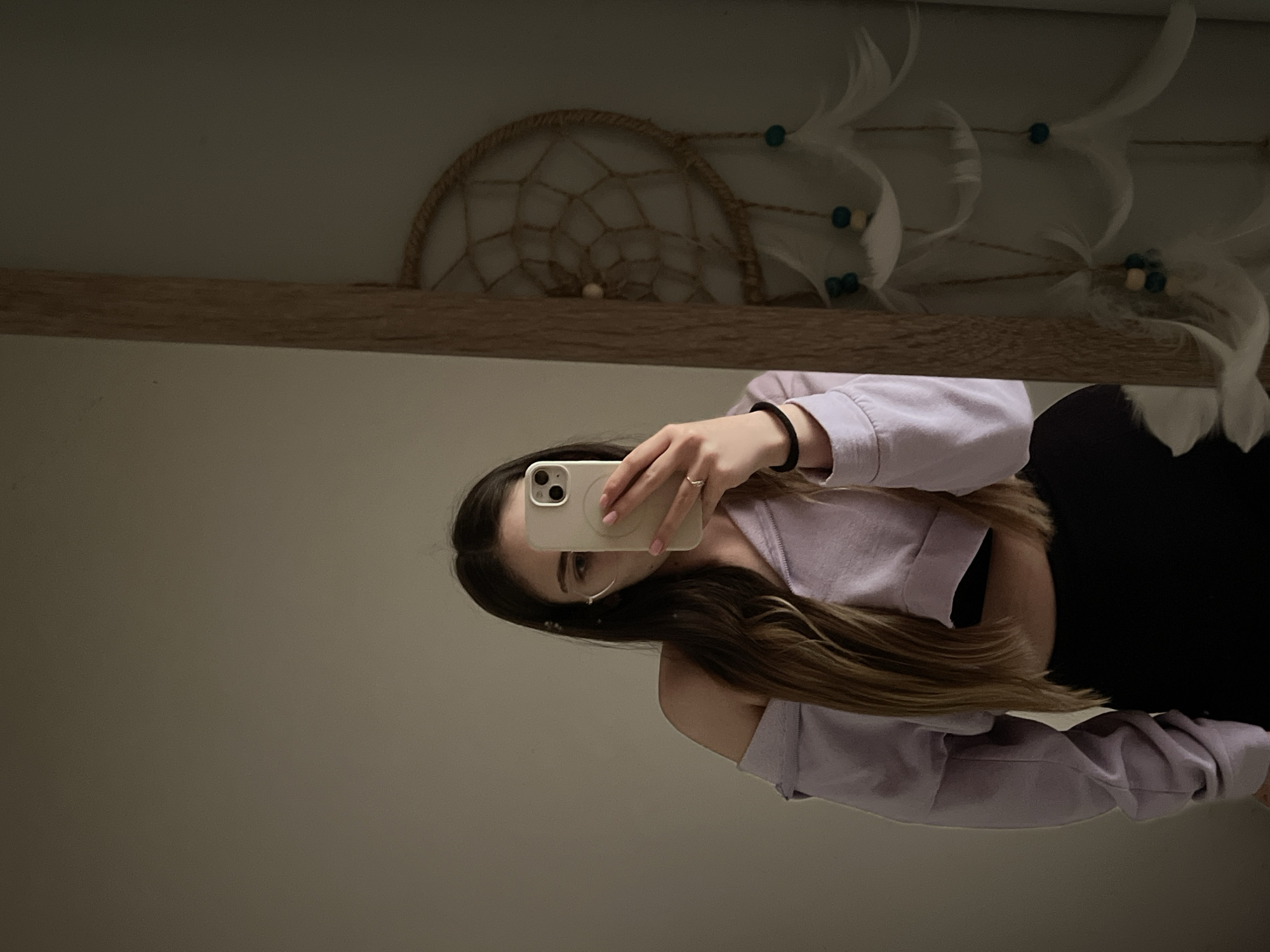 A person is holding a smartphone in front of a mirror, presumably taking a selfie. The individual is wearing a lavender-colored top, with a dark garment visible underneath. The mirror's frame is made of wood, and a dreamcatcher is positioned on a shelf behind the mirror. The environment appears to be an indoor setting. The city is Unknown, Unknown.