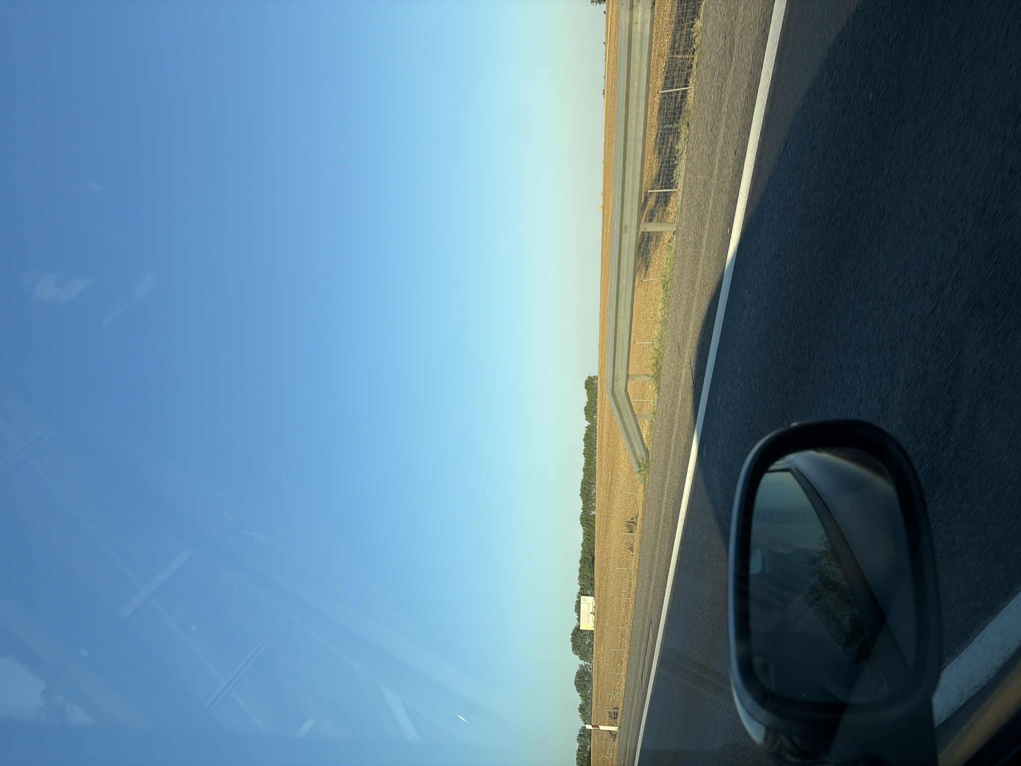 An exterior, eye-level, landscape shot shows a bright blue sky above a rural scene taken from a moving vehicle. In the foreground, the dark asphalt of a highway fills the bottom of the frame, with a white line marking the lane. To the left, a car's side mirror shows a reflection of trees and the sky. In the midground, a dry, tan-colored field stretches towards a line of green trees in the distance. A metal guardrail runs along the edge of the field, separating it from the highway. Behind the field and trees, a white building with a sign that reads "PREDALVINA" is visible. The scene is located near Báb, Slovakia.