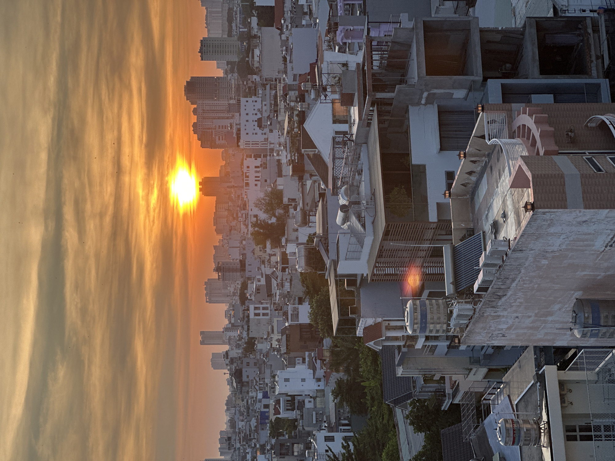 The image depicts a sunset over Ho Chi Minh City, Vietnam. In the foreground, there are densely packed buildings with a variety of architectural styles. Some buildings have water tanks on their roofs. In the background, the skyline is visible with taller buildings. The sun is setting, casting a bright orange glow on the sky and reflecting off the buildings.