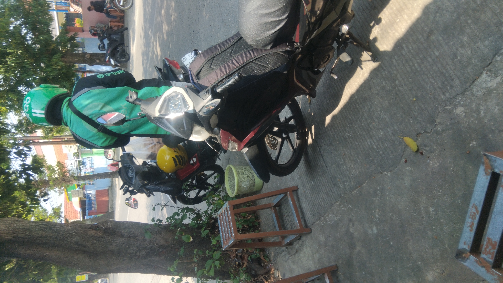 A man wearing a green Gojek uniform and helmet stands next to a motorcycle on a paved street in Proyonangg, Indonesia. The man is facing away from the camera, with his back to the viewer. The motorcycle is black with red accents and has a backpack attached to the back. Another motorcycle, also black, is parked behind the first one. In the background, there are trees, buildings, and other motorcycles. The image appears to be taken in the daytime, with sunlight casting shadows on the ground.