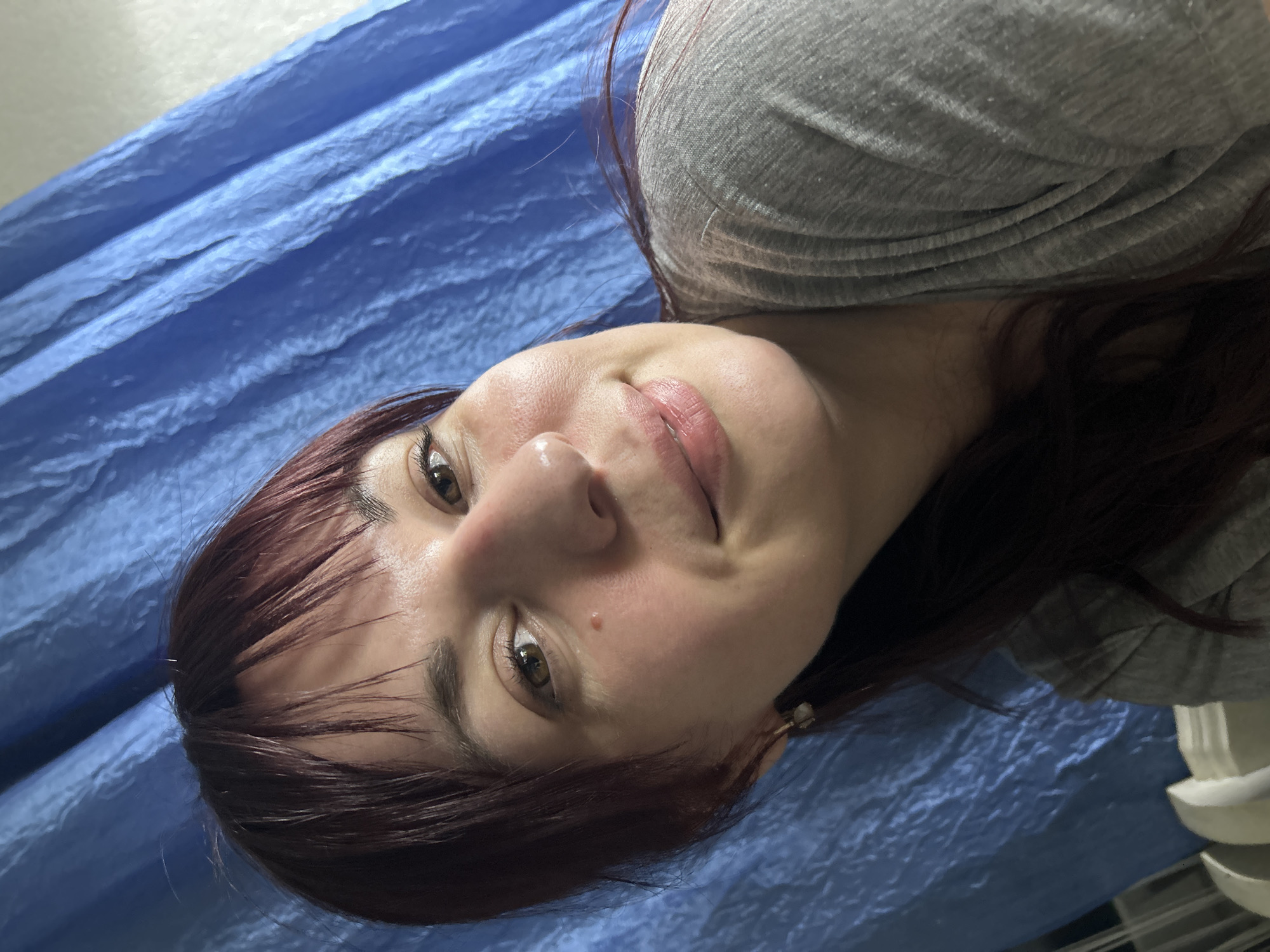 A woman with reddish-brown hair and fair skin is lying on a blue, creased surface, possibly a tarp. She is wearing a gray t-shirt. Her head is slightly tilted, and she is looking at the camera with a faint smile. A small mole is visible on her cheek. The environment appears to be indoors. There is no evidence to suggest this image was taken in Фоки, Russia.
