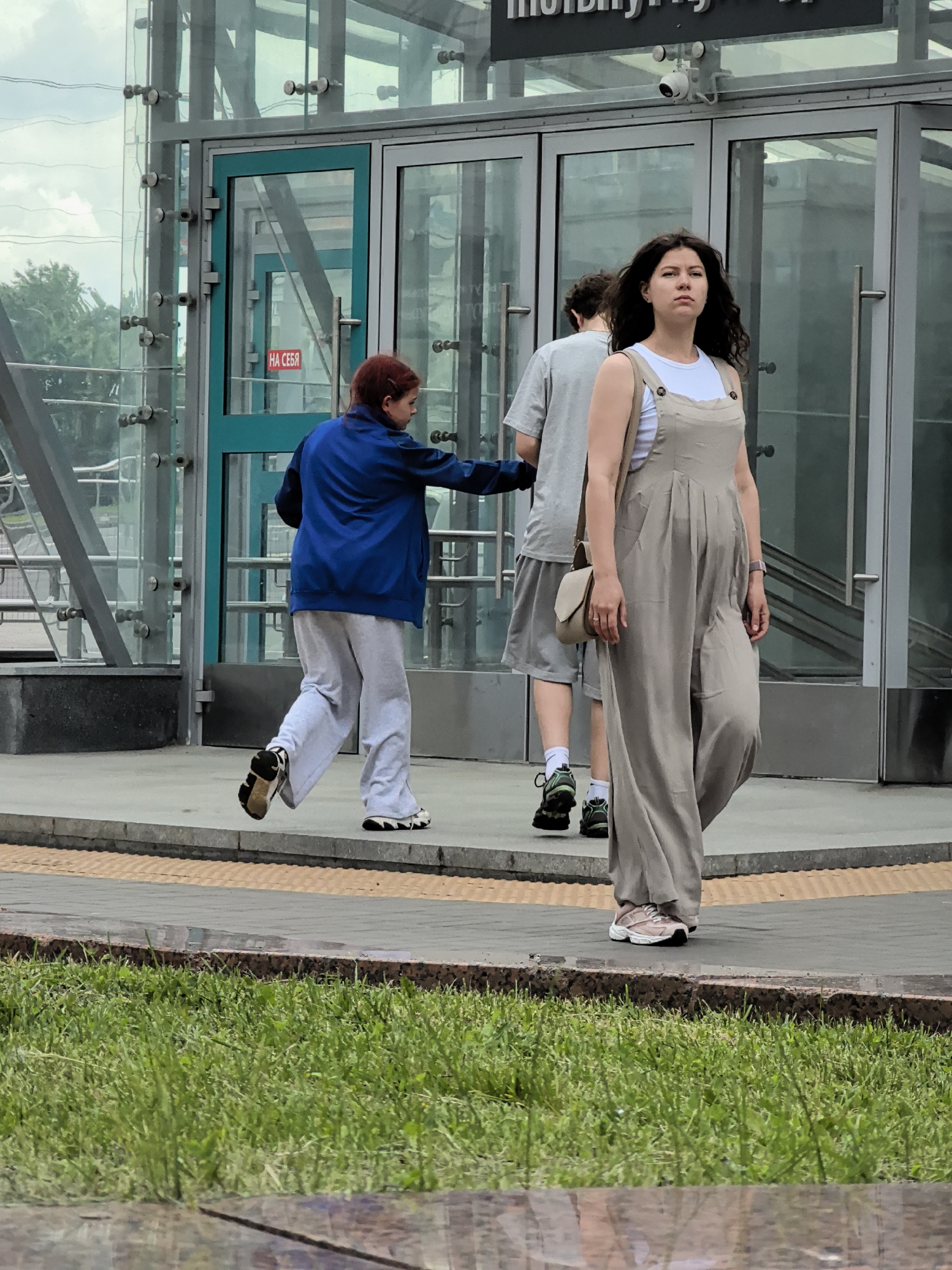 A medium shot captures three individuals near a glass-enclosed structure with large glass doors and metal framing. In the foreground, a woman with dark, wavy hair walks forward, wearing a light beige jumpsuit over a white tank top and a small crossbody bag. She has on pink and white sneakers. Behind her and to the left, a person in a blue jacket and light grey sweatpants with athletic shoes is captured in motion, appearing to be walking or running. Further back and to the right, another individual in a grey t-shirt and shorts stands, wearing dark athletic shoes. The environment appears to be an urban outdoor setting, with a sidewalk, grass, and trees visible in the background. A security camera is mounted above the glass doors.