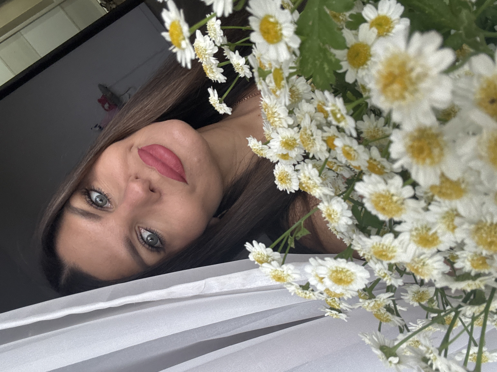 A woman with long brown hair and blue eyes is partially visible, holding a bouquet of white daisies with yellow centers. She is wearing pink lipstick and a light-colored necklace. A white curtain is on the left side of the frame, and a dark background is behind her. The scene appears to be indoors. The image does not contain any specific geographic markers that would indicate it was taken in Chaikovsky, Russia.