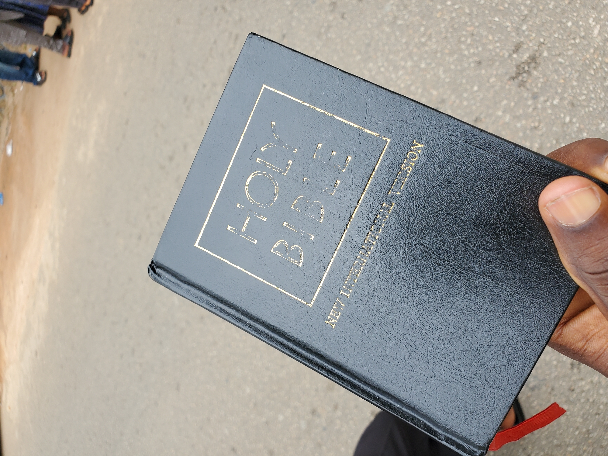 A person holds a black, leather-bound "Holy Bible, New International Version" in their right hand. The photo is taken outdoors, likely in a public area in Jos, Nigeria. The backdrop is a grey concrete surface where other people are visible in the background.