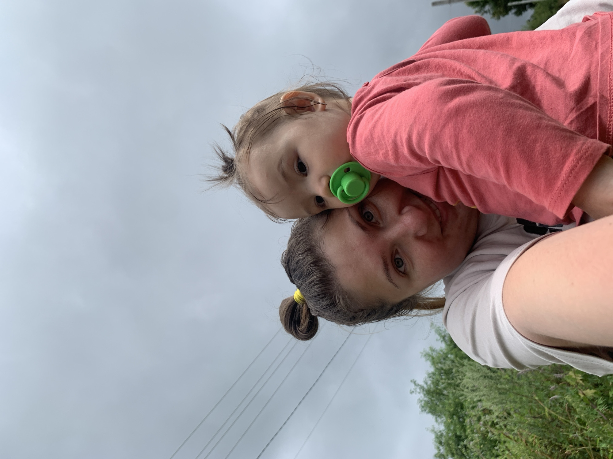 A selfie is taken by a young woman who is holding a baby. The woman is in the foreground and is smiling at the camera. The baby is held in her arms and is looking away from the camera. The baby has a pacifier in its mouth. The background is a cloudy sky and some green foliage. The scene appears to be outdoors, possibly in a rural or suburban area. There are power lines visible in the upper left corner of the image. The image does not provide specific details to determine the exact location within Zasechnyi, Russia, but it appears to be an outdoor setting.