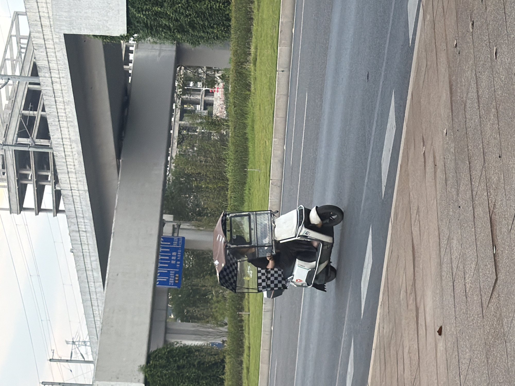 A person is riding a white, three-wheeled electric scooter with a canopy on a paved road in the city of Jiangjin, China. The scooter has a clear plastic windshield and a dark checkered canopy. The person is wearing dark clothing and appears to be looking down at something in their hands. The scooter is in motion, heading away from the camera. The road is flanked by a grassy median and a sidewalk made of textured concrete tiles. In the background, an elevated highway structure is visible, along with a partially constructed building and lush green trees. Overhead power lines and utility poles are also present. A blue traffic sign with white text is mounted on the underside of the elevated highway. The sky is overcast.
