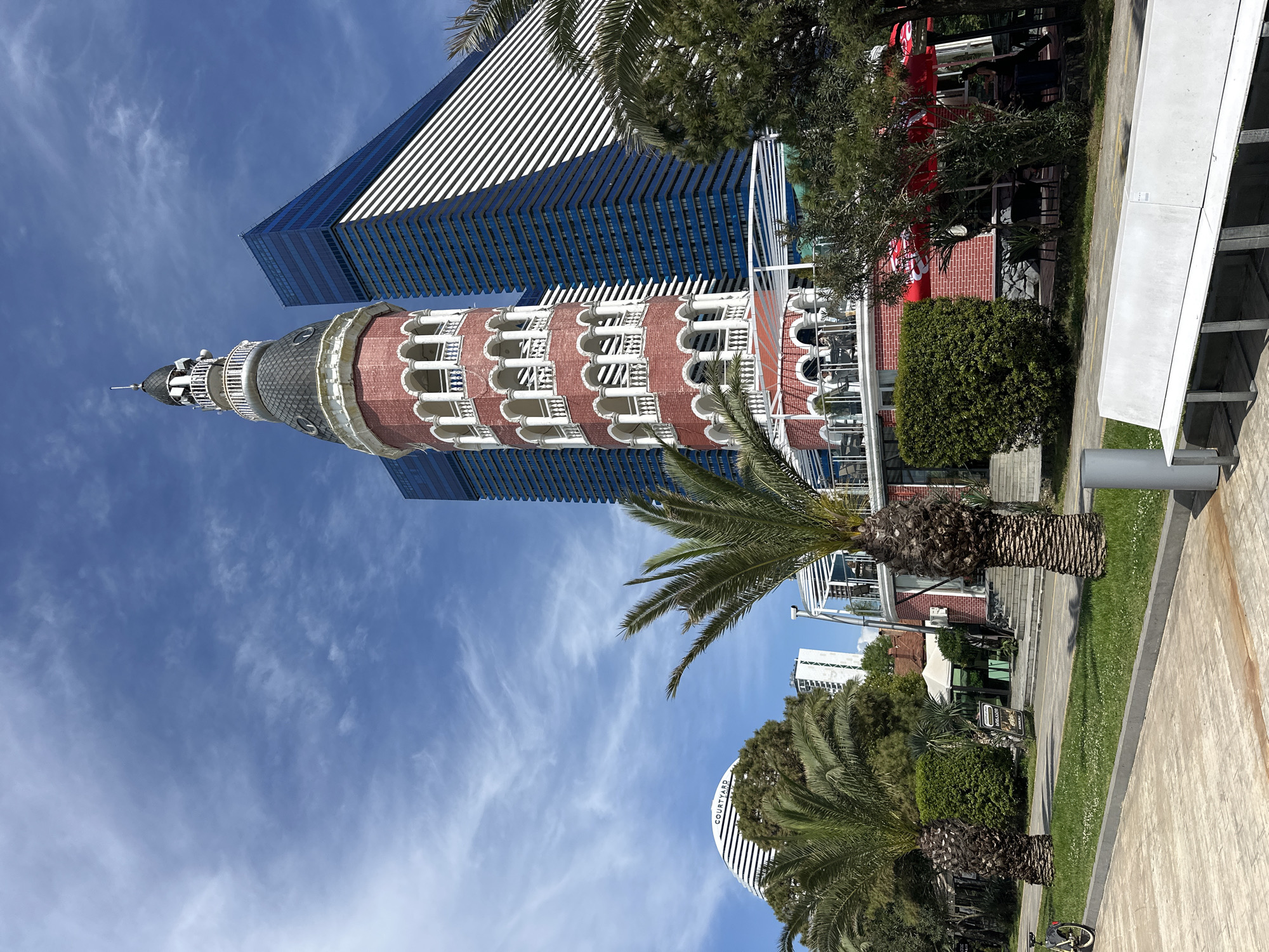 The image captures the Chacha Clock Tower in Batumi, Georgia, juxtaposed against a modern skyscraper with blue-tinted windows. The tower is characterized by its red brick construction, white balustrades, and a dark, pointed roof with a spire. Palm trees are in the foreground, adding to the subtropical atmosphere. To the right of the tower is part of a brick building with a visible Coca-Cola advertisement. A portion of a concrete structure is also visible on the right side of the image. The sky is blue with scattered clouds.