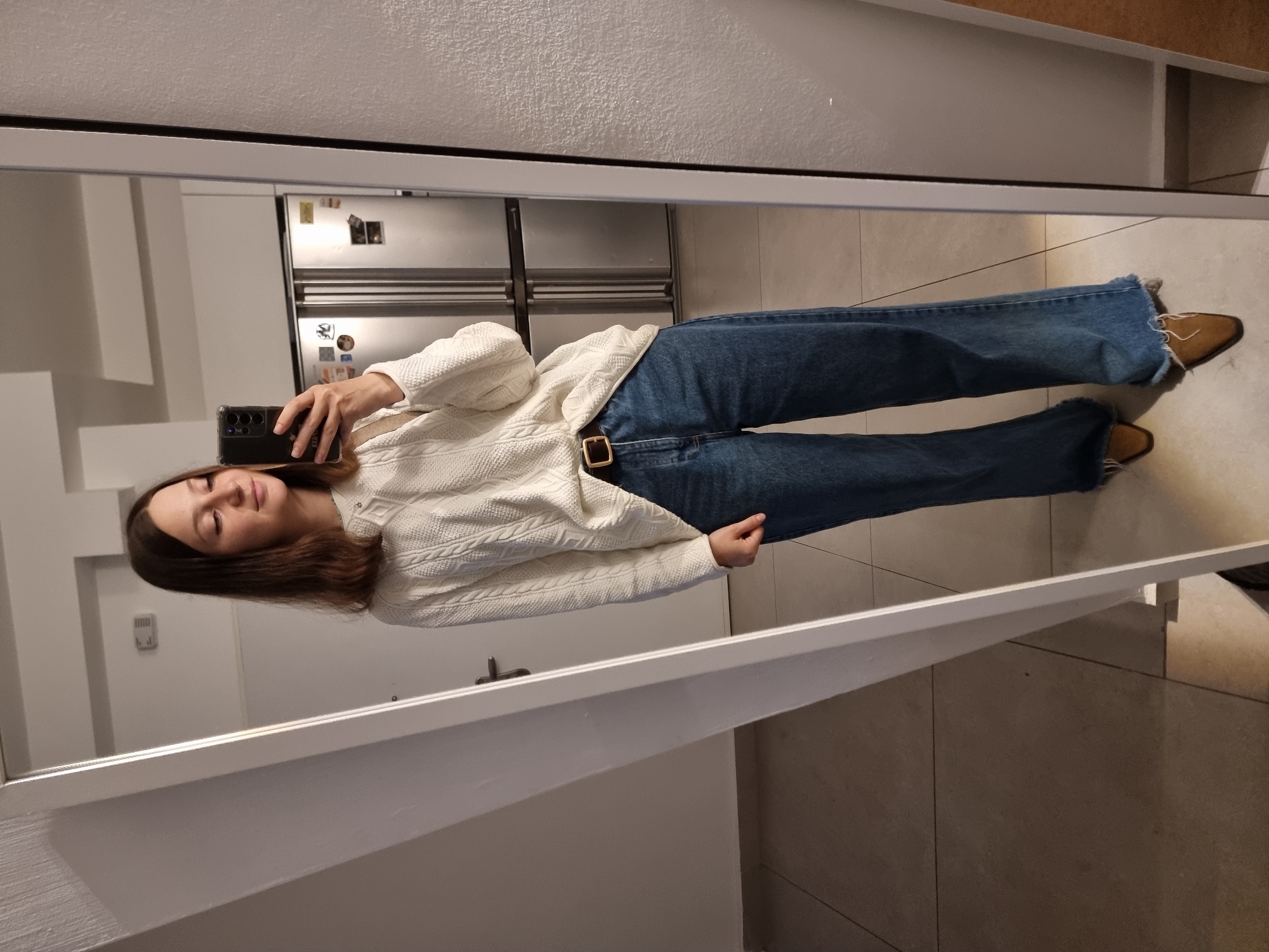 A woman is taking a selfie in a mirror. She is wearing a white cable knit sweater, blue jeans with a brown belt, and tan pointed-toe boots. The location appears to be an apartment with light-colored tile flooring and a modern kitchen with a stainless steel refrigerator in the background. The location is in Ramat Gan, Israel.