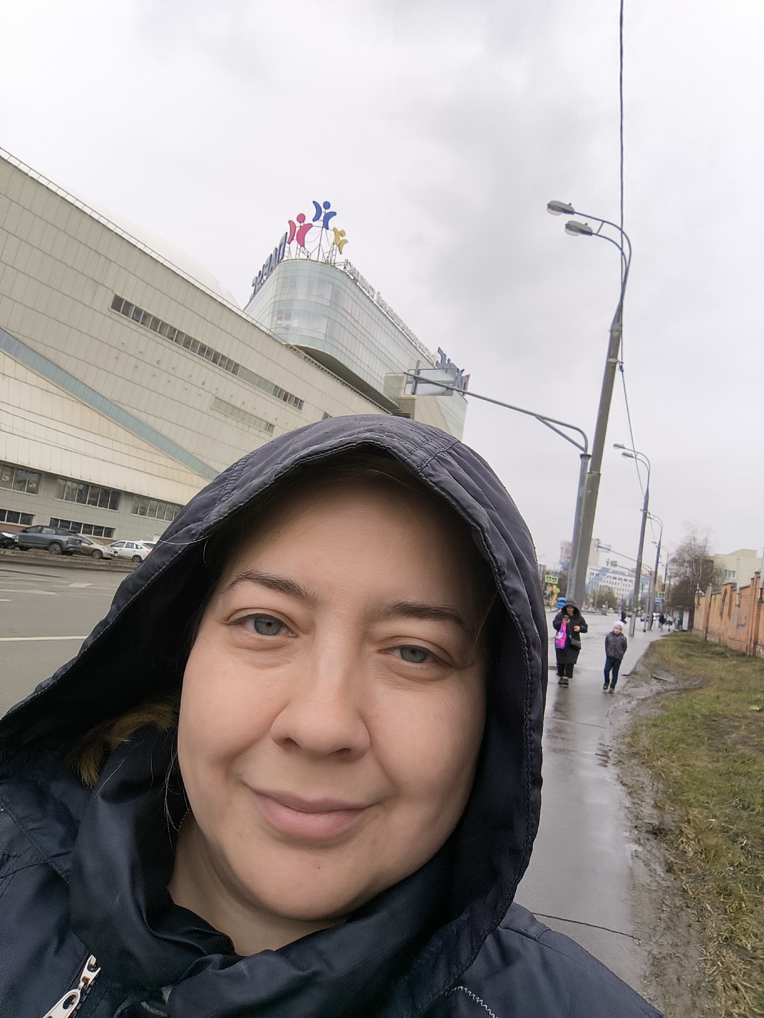 A selfie taken outdoors on a cloudy day shows a woman in a dark hooded jacket. The woman faces the camera with a slight smile. Behind her, a large, modern building with a multi-colored logo is visible. People are walking on the sidewalk. Streetlights line the street. Cars are parked on the opposite side of the street. The ground is wet. The city is Unknown, Unknown.