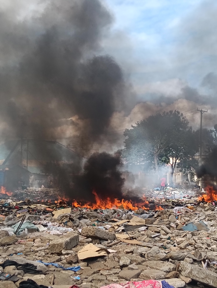 This image depicts an outdoor scene in an unidentified area within the city of Nairobi, Kenya, characterized by widespread debris and active fires. The foreground consists of an uneven ground heavily covered with concrete rubble, various refuse, and discarded textiles in multiple colors. Across the midground, several distinct lines of bright orange flames are actively consuming this debris, generating voluminous plumes of dense, black smoke that ascend into the sky and spread horizontally, obscuring significant portions of the background.
A lone individual, clad in a red garment, is partially visible on the right side of the midground, positioned amidst the smoke and debris. Their specific actions are not discernible beyond their presence in the scene. In the background, a green-leafed tree and a wooden utility pole with overhead wires are visible through the smoke on the right. Indistinct structures are faintly discernible through the haze on the left. The sky above shows patches of blue amidst an overcast and heavily smoke-filled atmosphere. No direct interaction between the visible individual and the fire is clearly observable.