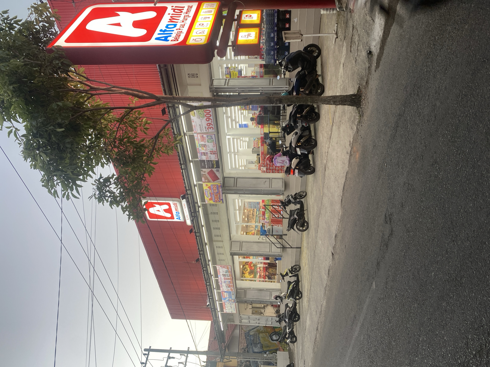 The image shows a street scene in Bantaeng, Indonesia, with a brightly lit Alfamidi convenience store as the central focus. The store has a large red facade and a prominent red and white Alfamidi sign. Parked motorcycles line the sidewalk in front of the store, and several people are visible inside and near the entrance, suggesting it is open for business.
The street in the foreground is asphalt, with a concrete sidewalk bordering the store. Overhead power lines crisscross the sky, and a tree with green foliage grows in front of the Alfamidi sign. A truck with graffiti on its side is partially visible on the left side of the image. The overall impression is a typical urban commercial setting in Indonesia, likely during the late afternoon or early evening, given the lighting.