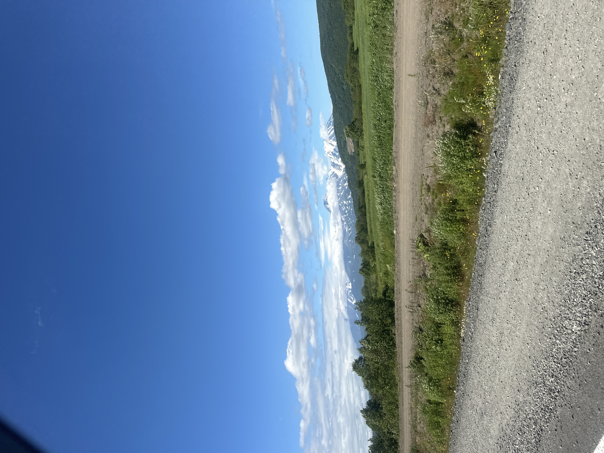 The image displays a scenic natural landscape under a bright blue sky with scattered white clouds. In the foreground, a gravel road leads into the frame from the lower left corner, bordered by green vegetation and some wildflowers. Beyond the road, a line of trees forms a transition to a lush green meadow. In the background, snow-capped mountains rise majestically, with some of the peaks obscured by clouds. The overall impression is one of a clear, sunny day in a mountainous region.
The image is located in the vicinity of Petropavlovsk-Kamchatsky, Russia. The specific scene depicts a view towards the Avachinsky and Koryaksky volcanoes, which are prominent landmarks near the city. The foreground features a gravel road and surrounding vegetation, characteristic of the rural and natural areas surrounding Petropavlovsk-Kamchatsky. The distant mountains with snow caps are consistent with the volcanic landscape of the Kamchatka Peninsula.