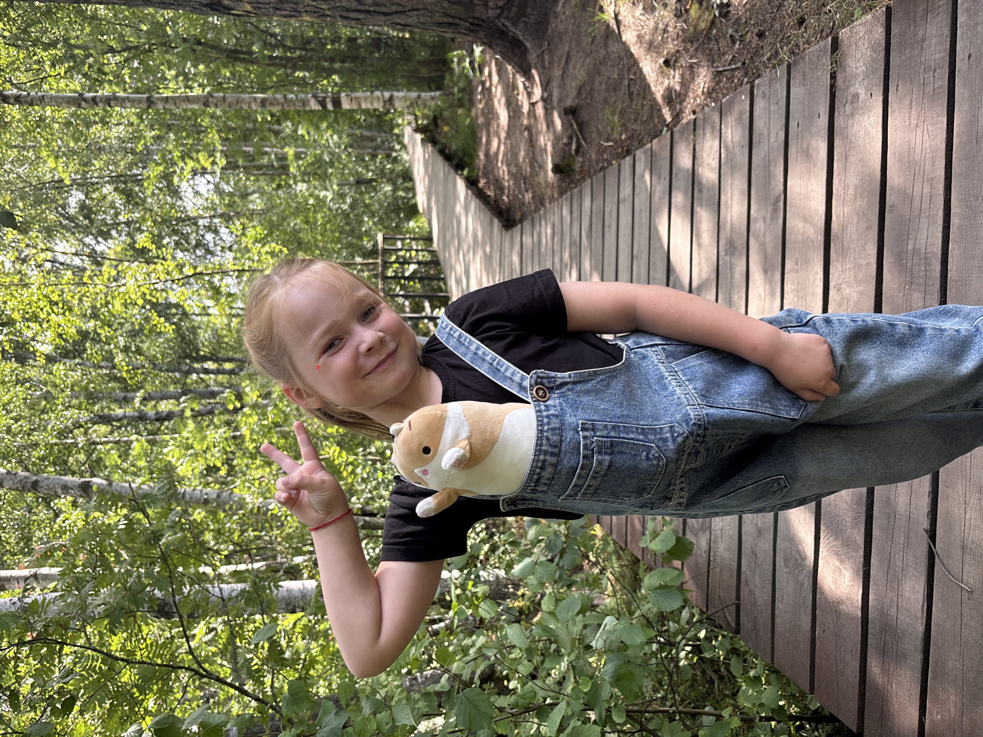 A young girl with blonde hair tied back and makeup on her cheeks is standing on a wooden boardwalk in a forest. She is wearing a black t-shirt and denim overalls. She is holding a stuffed toy hamster in her overalls pocket and making a peace sign with her left hand. The boardwalk is made of weathered wooden planks and extends into a forest with birch and pine trees. The foliage is dense and green, indicating a summer setting. Sunlight filters through the trees, casting dappled shadows on the boardwalk. The location is identified as Beloostrov, Russia.