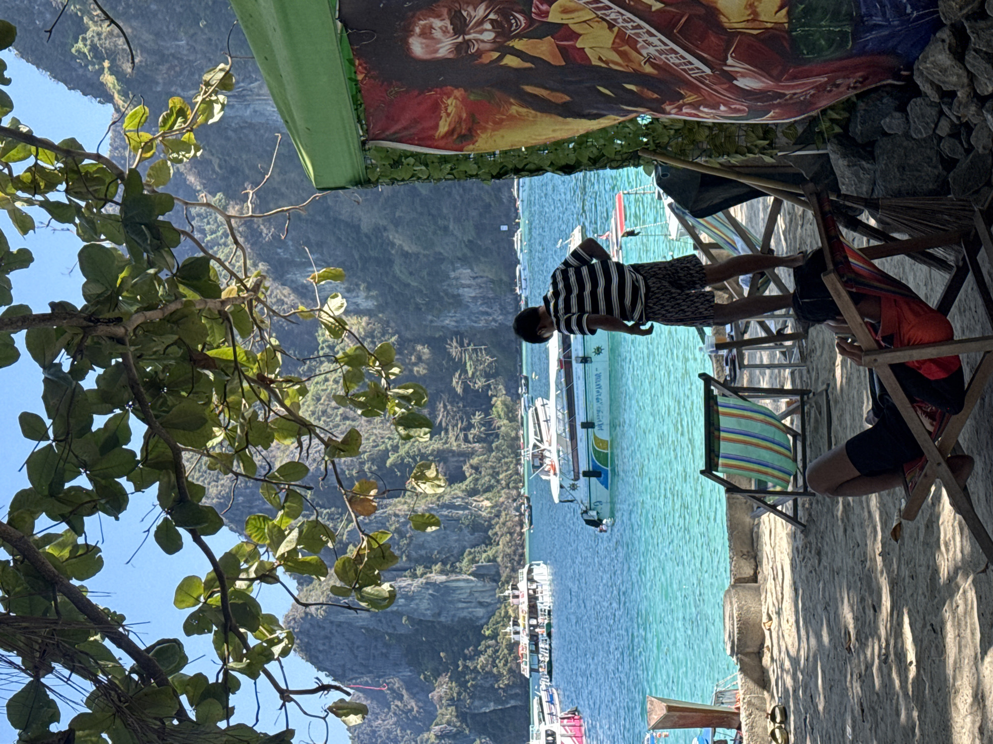 Here's a description of the scene:
The image captures a coastal scene in Ao Nang, Thailand. Turquoise water fills the majority of the frame, speckled with numerous boats. A lush, mountainous coastline serves as a scenic backdrop. Closer to the foreground, there are individuals and elements of a beachside establishment. One person is standing, wearing a striped shirt and patterned shorts. Another person is seated in a beach chair with shorts, with a red cloth draped on the chair. A variety of beach chairs are lined up. Overhanging the scene is a green awning and a banner with a portrait of Bob Marley, lending a relaxed, tropical vibe. A tree with dense green leaves frames the left side of the image. The ground appears to be a mixture of sand and concrete, with shadows cast by the overhead structures.