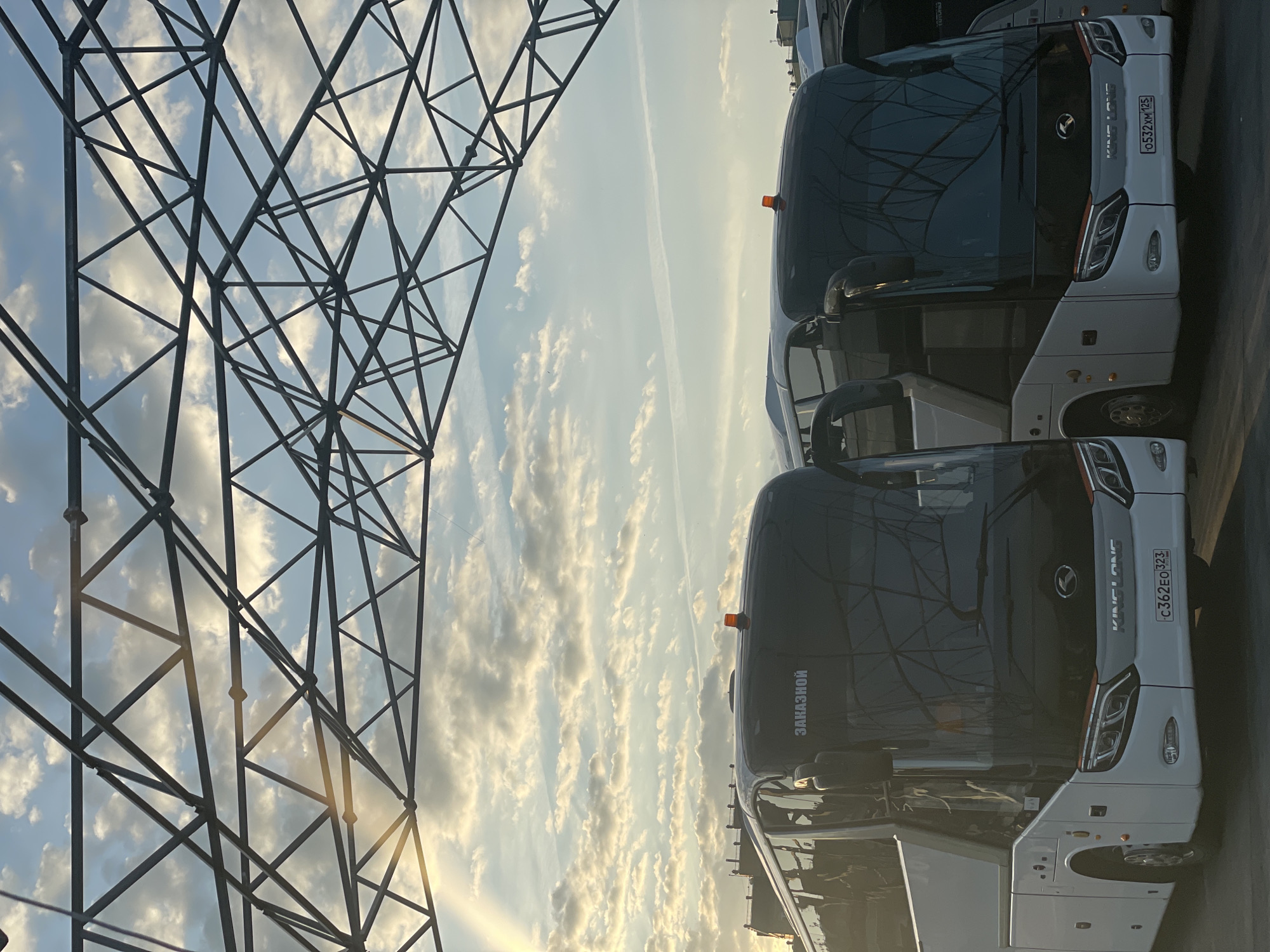 The image shows a fleet of white and black buses parked outdoors, likely in a depot or parking lot, under a large metal canopy structure. The sky above is partly cloudy with the sun breaking through, casting light rays and creating a warm, golden hue. The buses are modern with dark tinted windows and prominently display "KING LONG" branding on their fronts. One bus has "ЗАКАЗНОЙ" (meaning "custom" or "chartered" in Russian) written on its upper front panel. License plates are visible, with one reading "C362EO 323" and another "0532XM 125". The scene appears to be a typical view of a transportation hub or fleet parking area in Kazan, Russia, likely during the late afternoon or early morning given the quality of light. No people are visible in the frame.