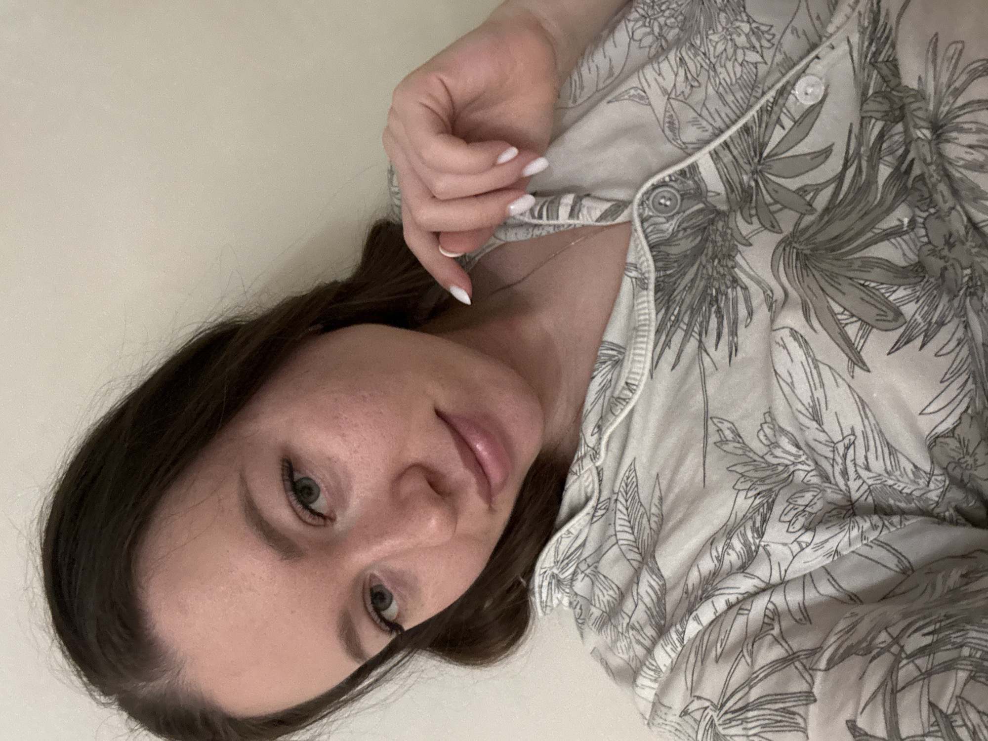 A woman with long brown hair and pale skin is looking at the camera. Her left hand, with white nail polish, is near her collarbone. She is wearing a button-down shirt with a white background and a grey botanical print. The background appears to be a plain, light-colored surface. The location is not discernible from the image.