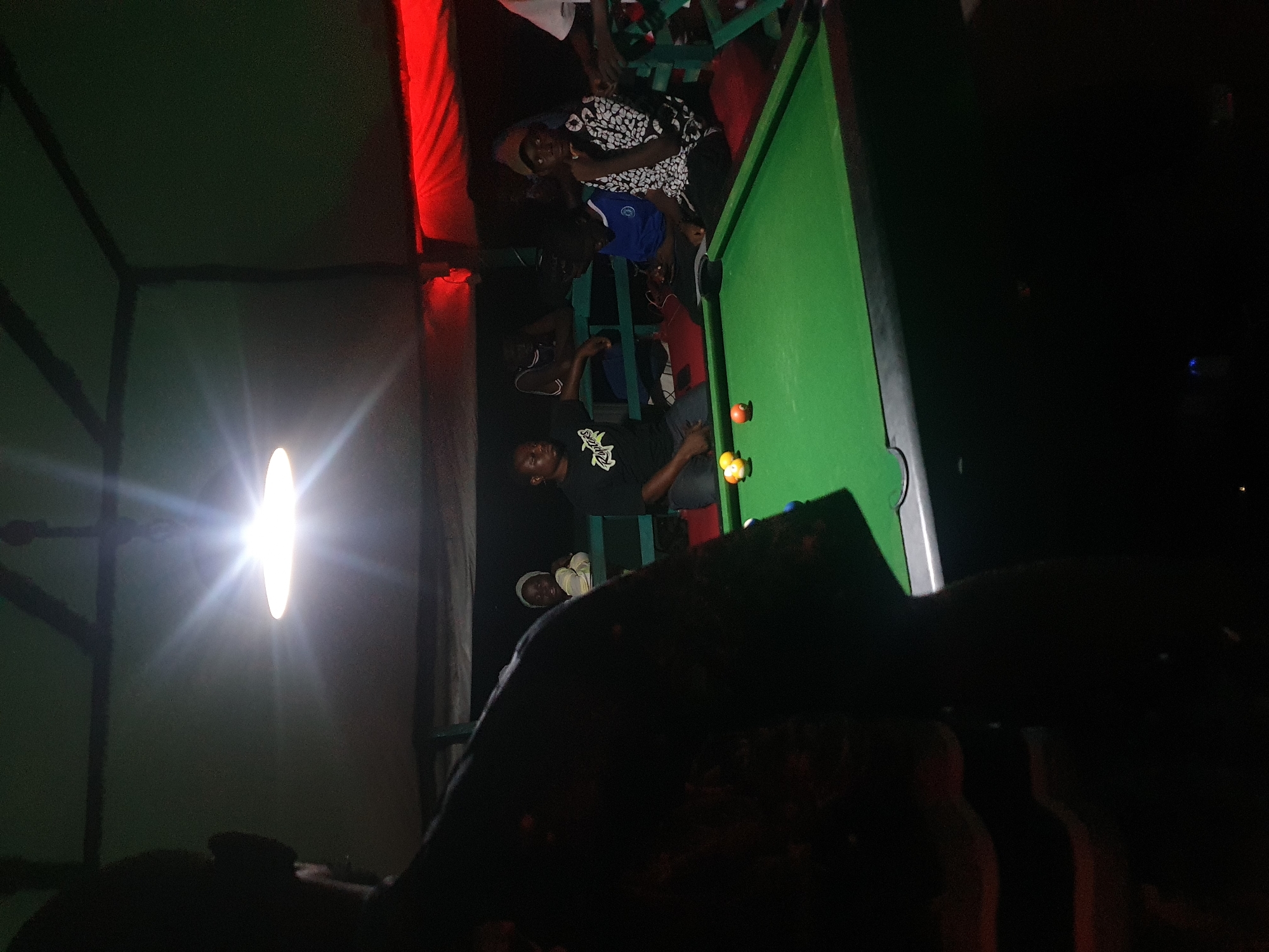 The image depicts a group of people gathered around a pool table in what appears to be an indoor or covered outdoor setting at night. The scene is dimly lit, with a bright light source, possibly a lamp, casting a strong glare. A pool table with green felt and several balls is visible, with some individuals positioned as if playing or observing the game. One person is wearing a black t-shirt with white text that is partially obscured but appears to be a graphic design or logo. Another person is wearing a blue shirt, and others are in casual attire. A red fabric is visible in the background, adding a splash of color to the dark environment. The overall atmosphere suggests a casual social gathering. The mention of Lokoja, Nigeria provides geographical context for the scene.