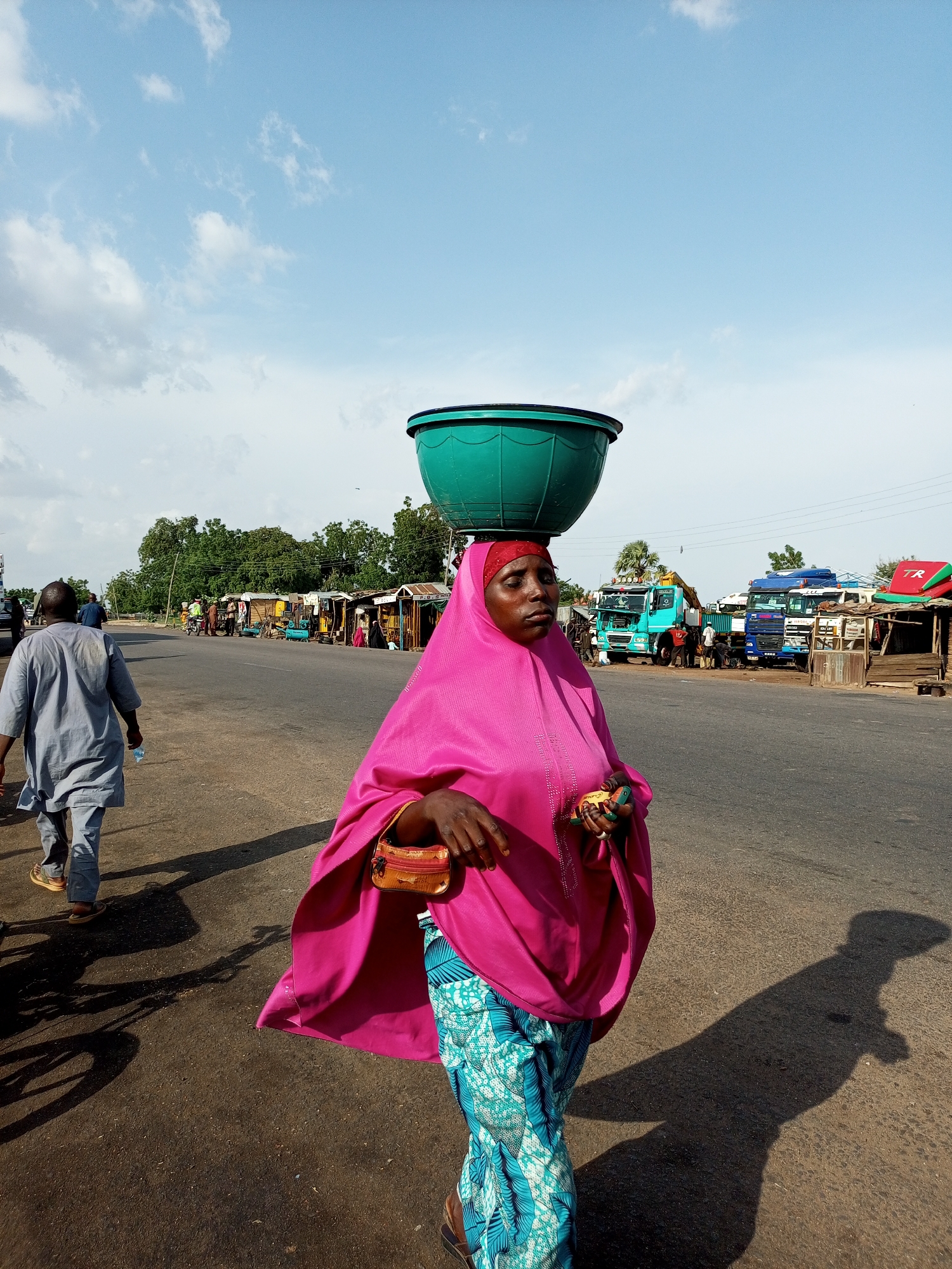 A woman with a large green bowl balanced on her head walks on the right side of a paved road in Gadao, Nigeria. She wears a pink hijab and a brightly patterned blue and turquoise skirt. A small, tan purse is clipped to her waist. Her right hand is raised, holding what appears to be a mobile phone. She casts a long shadow on the road. In the background, a man walks away from the camera on the left side of the road, carrying a water bottle. Across the road, a row of small market stalls and parked trucks are visible. The sky is blue with scattered white clouds.