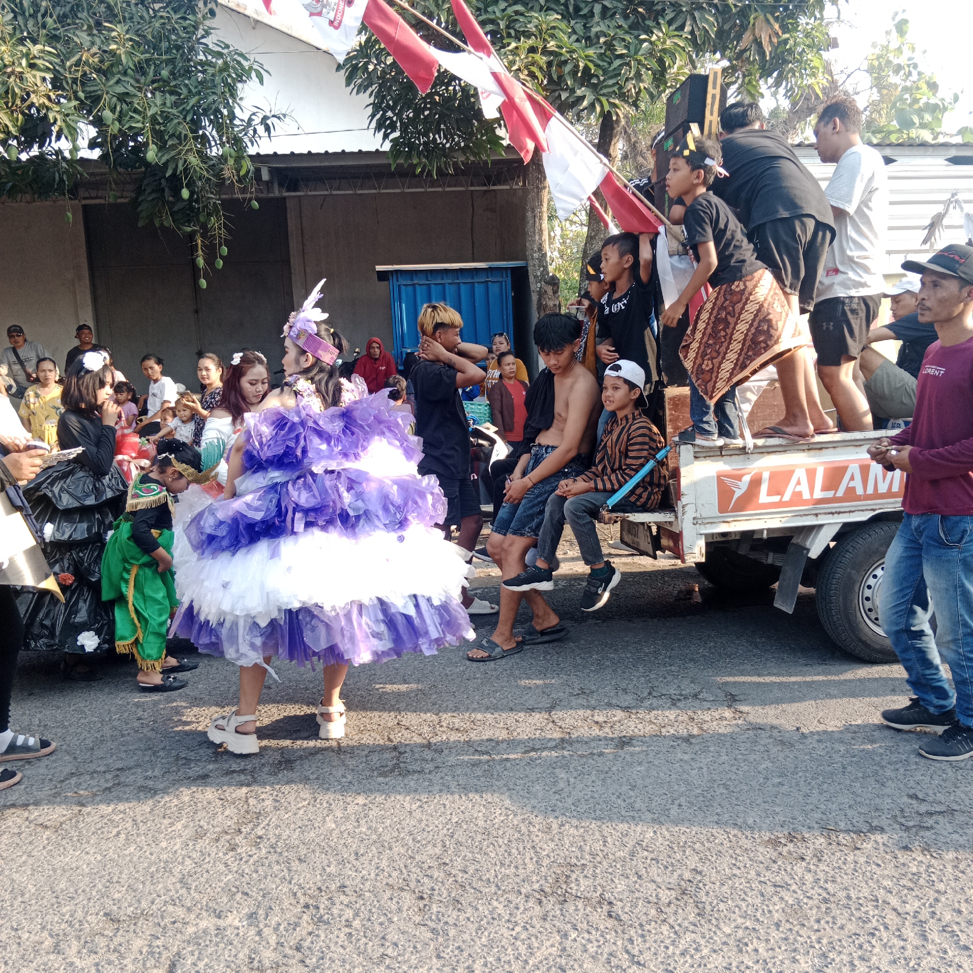 The image depicts a street scene in what appears to be a procession or parade in Tegalrejo, Indonesia. In the foreground, a young girl wearing a large, layered purple and white dress with a decorative headband is positioned prominently. To her left, another young girl in a black costume with a ruffled skirt and a white flower adornment is also visible. A smaller child in a green costume with gold trim stands between them.
In the background, a white truck serves as a platform for several individuals. Two young boys are seated on the edge of the truck, one wearing a striped shirt and a white cap, the other with his torso bare and wearing denim shorts. Other people are standing or sitting on the truck, some holding Indonesian flags. The truck itself has the logo "LALAM" in orange letters.
The environment suggests a sunny outdoor setting. Trees with green leaves and mangoes are visible overhead. A building with a gray facade is behind the people. The ground is paved with asphalt. Several adults are scattered throughout the scene, some watching the procession and others participating. The overall atmosphere appears festive.